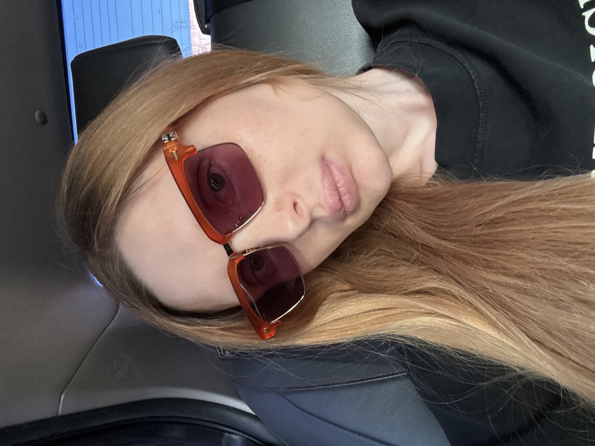 This is a portrait of a young woman with long, blonde hair, wearing orange-framed sunglasses with dark lenses. She is lying down, with her head tilted to the side, and her face is partially obscured by her hair and sunglasses. Her lips are slightly parted. She is wearing a black sweatshirt.
The background suggests she is inside a vehicle, possibly a car, as indicated by the seat texture and the glimpse of what appears to be a headrest. There is a hint of blue in the upper left, which could be a window or a screen.
The lighting suggests it is daytime. There are no explicit indicators of the weather. The overall mood is casual and relaxed. There is no visible text in the image. The setting appears to be Krasnodar, Russia, based on the provided location context.