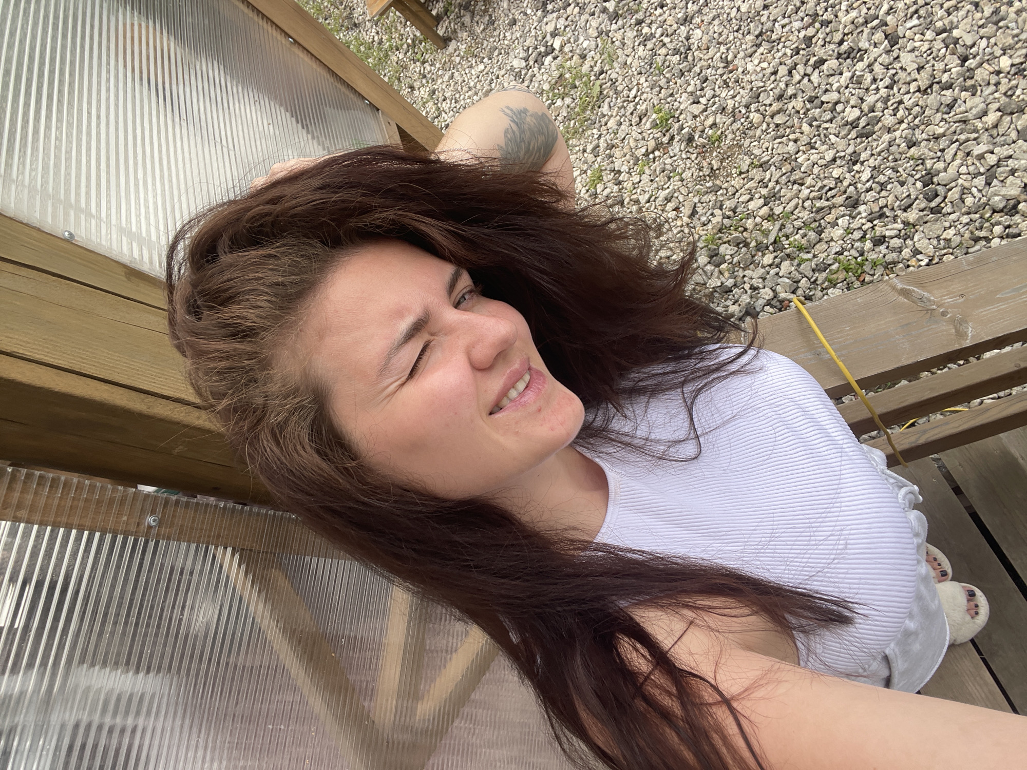The image shows a woman with long brown hair lying down, winking at the camera. She has a tattoo on her left arm and is wearing a white top. She appears to be lying on a wooden structure, possibly a bench or deck, located near a surface covered with gravel. The location is in the city of Мосметаллоконструкция Онт, Russia. There's a translucent, possibly plastic, panel behind her, suggesting she may be near or inside a greenhouse or similar structure. A yellow object is lying on the wooden structure nearby.