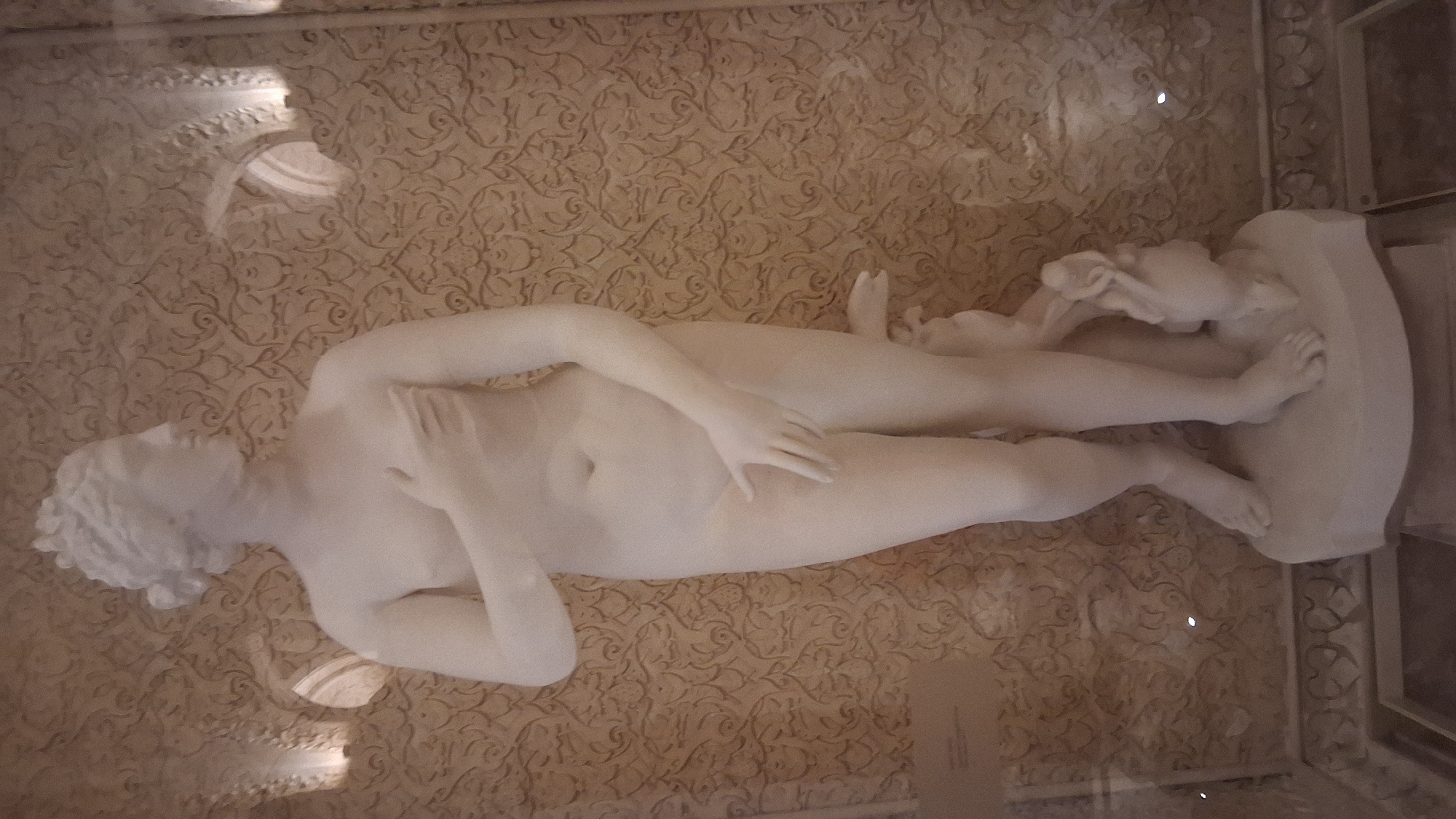 A white marble sculpture of Venus, the Roman goddess of love and beauty, stands against a patterned beige wall in the Pena Palace in Sintra, Portugal. The sculpture depicts Venus in a contrapposto pose, with her left leg slightly bent and her right leg supporting most of her weight. Her left hand is placed over her chest, and her right hand is gracefully positioned over her lower abdomen. She is depicted as nude, with her hair styled in an elaborate updo. At the base of the sculpture, a small figure, possibly Cupid, is visible. The wall behind the sculpture features an intricate floral or paisley pattern. The lighting suggests an indoor environment, likely a museum or gallery, with soft illumination highlighting the sculpture.