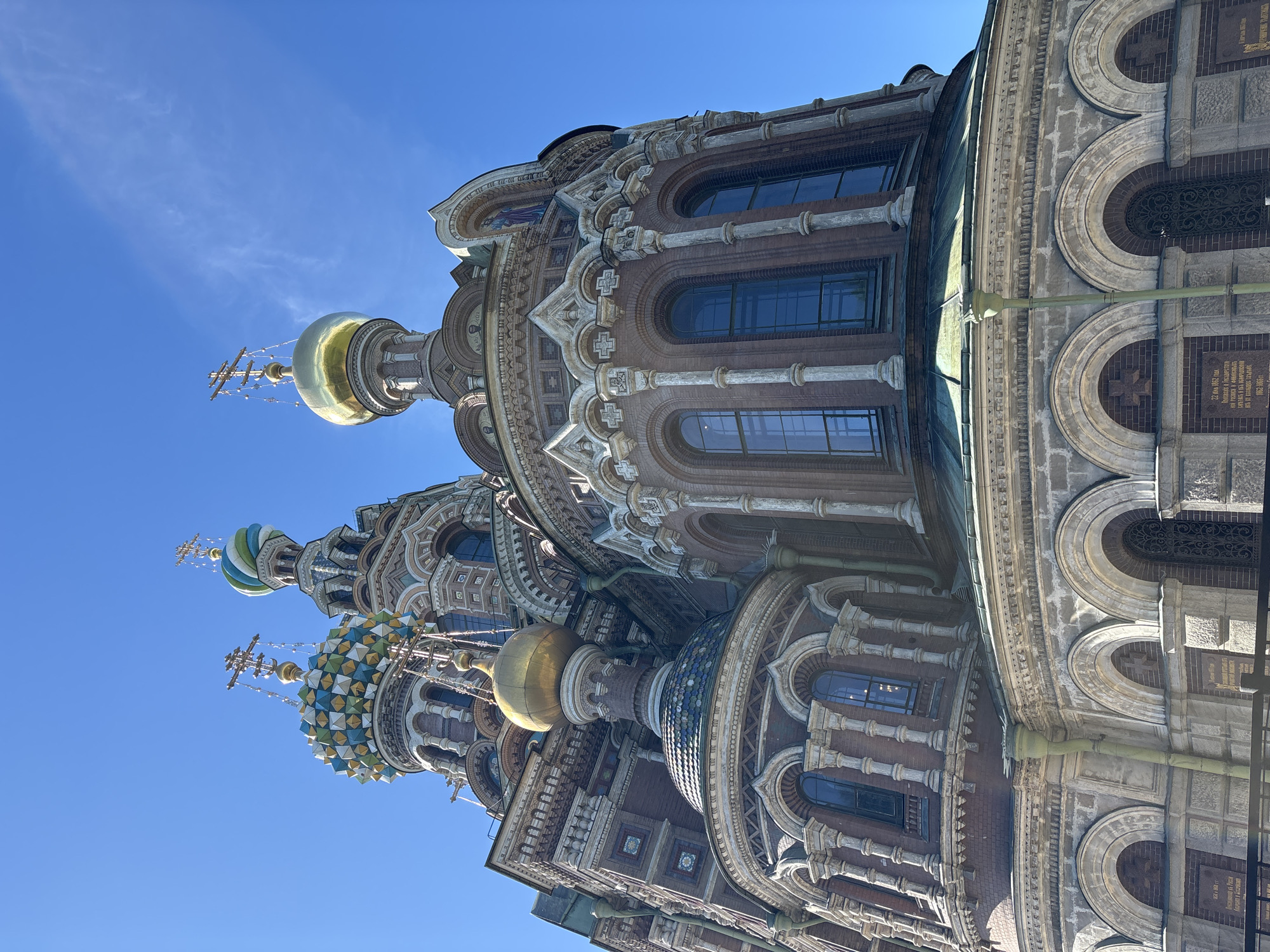 The image displays a detailed, low-angle exterior view of the Church of the Savior on Spilled Blood, located in Saint Petersburg, Russia, against a clear, vibrant blue sky with minimal wispy clouds. The ornate Neo-Russian architectural style of the church is prominent, featuring multiple domes. A large golden onion dome topped with an Orthodox cross is centrally positioned. To its left, another large dome exhibits an intricate geometric pattern of green, blue, and yellow tiles, also crowned with an Orthodox cross. A smaller, spiraling blue and white dome with a cross is visible towards the top right.
The church's facade is constructed from reddish-brown brick and decorative elements of lighter-colored stone or stucco. Numerous arched windows with dark or reflective glass are visible on the upper levels, framed by decorative columns and pilasters. Intricate friezes and cornices delineate various architectural tiers. Small mosaic icons or painted figures are present in arched niches on the upper sections. A smaller golden dome is visible in the lower-left portion of the structure. A light green downspout descends vertically along the mid-section of the church on the left side. The lowest visible section of the building features a base of rough-hewn light stone with arched recesses, below which are dark plaques with Cyrillic inscriptions, partially obscured by a black metal fence or railing at the very bottom of the frame. The scene is illuminated by bright daylight, casting subtle shadows. No individuals are present in the image.