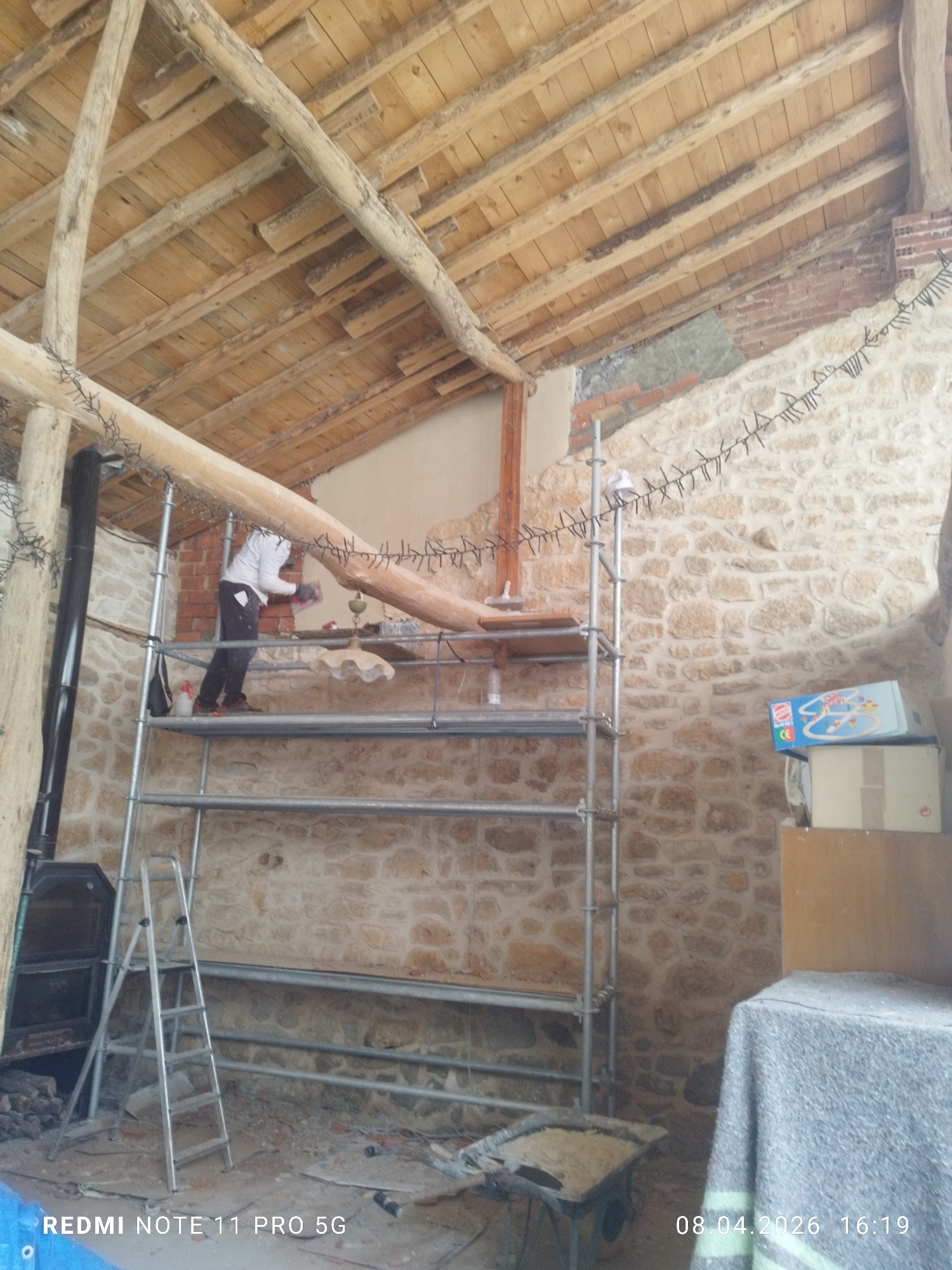 The image depicts the interior of a building in Caleruega, Spain, undergoing renovations. The structure features a wooden beam ceiling and stone walls. A man in white shirt and dark pants is standing on a metal scaffolding, seemingly working on the wall or ceiling. The scaffolding is positioned in the center of the frame. A ladder leans against the scaffolding. In the background, there's a wood stove. Several items are visible in the room, including a wheelbarrow containing an unknown substance, boxes, and furniture covered by a sheet. The environment suggests ongoing construction or restoration. A string of lights is draped along one of the wooden beams. The floor is littered with debris.