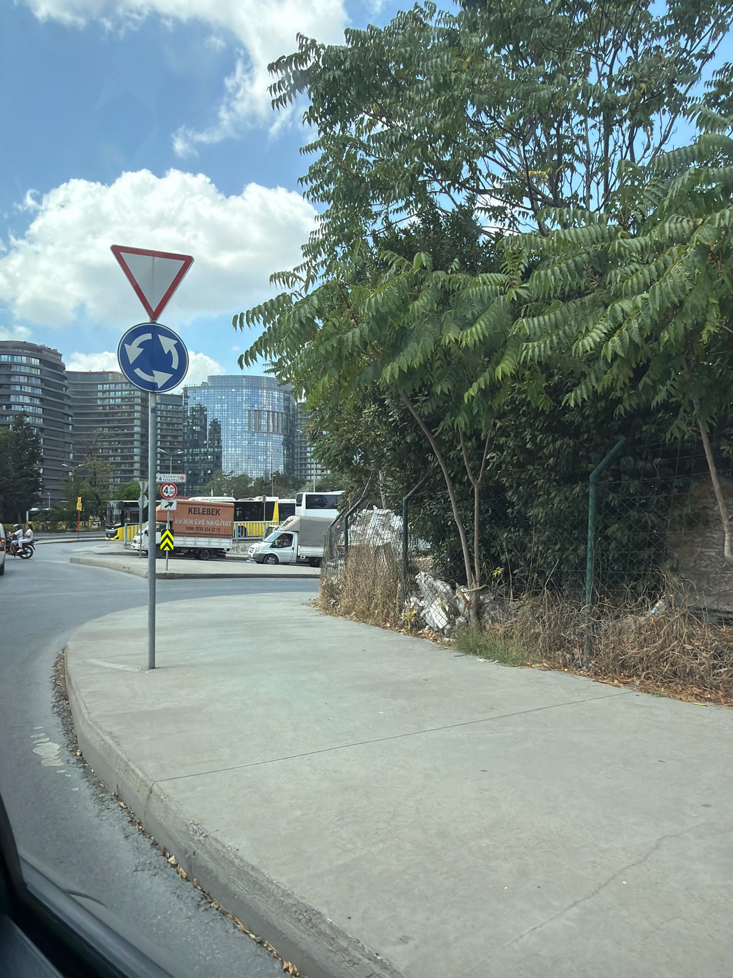 This image captures a street scene in Bakırköy, Turkey. In the foreground, a grey concrete sidewalk runs alongside a curved asphalt road. A grey metal pole supporting two traffic signs stands on the sidewalk. The upper sign is a red-bordered triangle pointing downwards, indicating "Give Way." Below it is a circular blue sign with two white arrows indicating traffic direction around a roundabout.
In the middle ground, several vehicles are visible. A yellow bus is stopped near the curb, and behind it, a white truck with an orange cargo box is parked. The text "KELEBEK EVDEN EVE NAKLİYAT" is visible on the truck's cargo box, along with a phone number. Another white truck with a grey cargo box is also present. A motorcycle with two passengers is seen in the left portion of the image.
The background is dominated by modern multi-story buildings with glass facades, suggesting an urban environment. Trees with lush green foliage are present on the right side of the image, partially obscuring the view of the buildings and creating a natural element within the cityscape. A chain-link fence is visible behind some of the trees, and there is some debris and dry vegetation along the fence line. The sky is blue with scattered white clouds. The overall impression is of a sunny day in a bustling city.