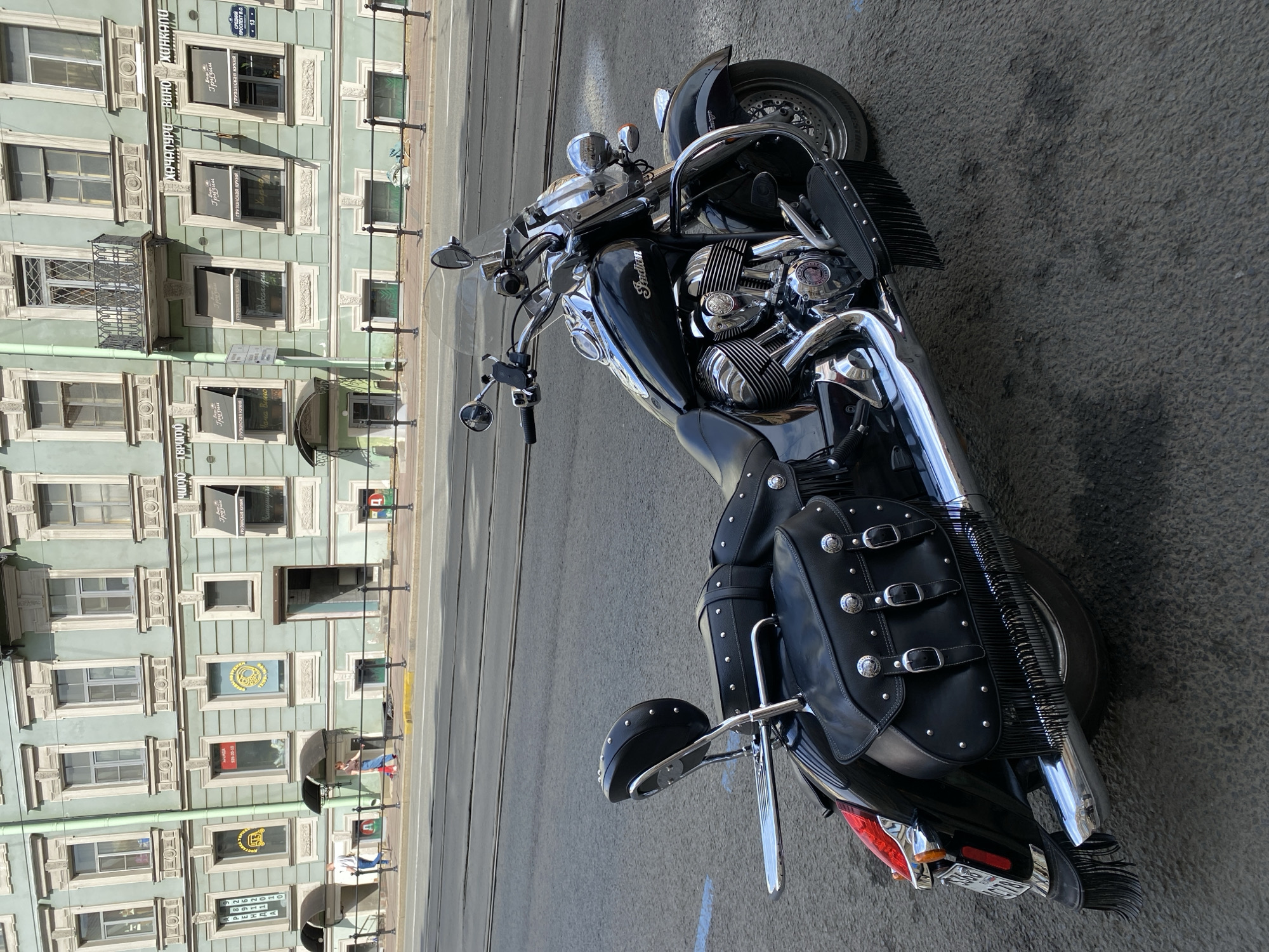A black Indian motorcycle is parked on a street in front of a light green building in Saint Petersburg, Russia. The building has multiple windows, some with awnings, and signs for businesses written in Cyrillic script. The street has tram tracks. The motorcycle has leather saddlebags with fringe, a passenger backrest, and chrome accents. The scene is captured from a low angle, looking up towards the building.