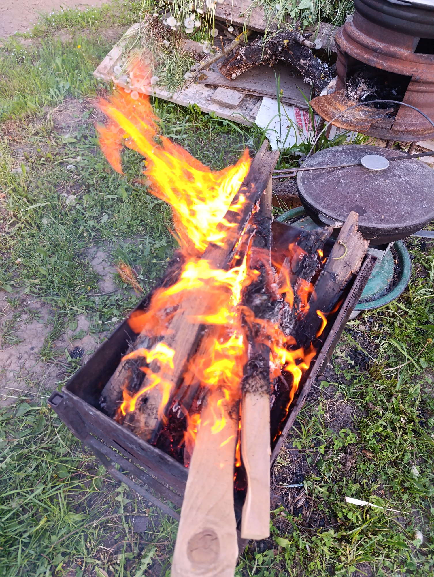 The image shows a metal fire pit containing burning wood. The flames are bright orange and yellow, rising high above the pit. The pit is situated on a patch of green grass in an outdoor setting. A cast iron pot with a lid rests near the fire pit. In the background, there is a wood pile and an old stove.
