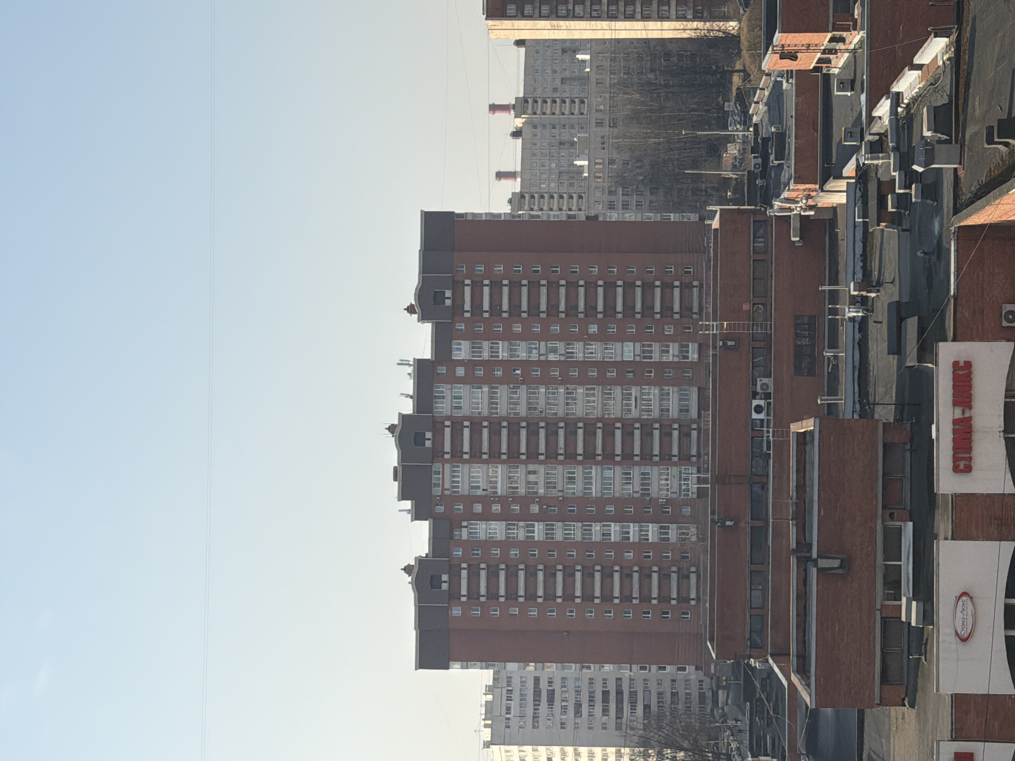 An elevated, long-range photograph depicts a dense urban landscape in Saint Petersburg, Russia, specifically oriented towards the prominent residential building at **Grazhdanskiy Prospekt, 76**. The sky is clear and light blue, with two faint, thin horizontal lines, possibly power cables, traversing the upper portion of the frame.
In the immediate foreground, a collection of dark, flat rooftops are visible, featuring numerous vents, pipes, and mounted air conditioning units. These rooftops belong to low-rise brick buildings. One such building displays a partial white facade with a red sign clearly reading "СТОМА-ЛЮКС" (STOMA-LUX), indicating a dental clinic. An external metal ladder is affixed to one of these brick structures.
The midground is dominated by a substantial multi-story residential building constructed primarily of red brick, with alternating sections of white balconies and windows. This building features a distinctive dark gray, segmented roofline. To its left, a segment of a street or pathway is visible, lined with bare deciduous trees, where very faint human figures and parked vehicles can be discerned.
The background encompasses a vast expanse of additional residential buildings, varying in height and architectural style, predominantly light gray or white, consistent with Soviet-era apartment blocks. Two prominent red and white striped industrial chimneys are visible in the far distance, rising above the residential structures. No explicit human actions or interactions are discernible due to the distance and scale. The scene is illuminated by natural daylight, casting subtle shadows.