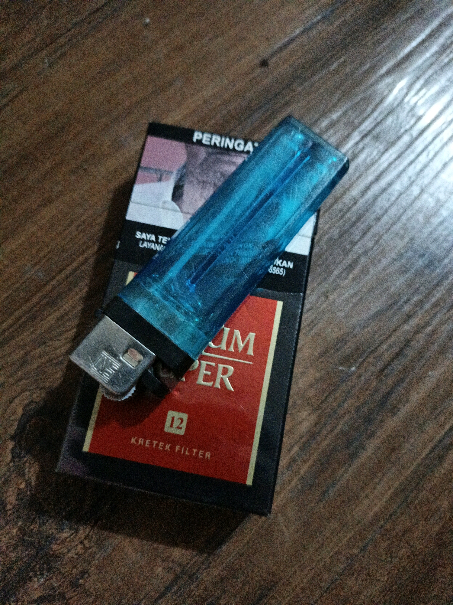 A blue lighter rests diagonally on a pack of cigarettes. The cigarettes are black with red and yellow branding. Text on the pack includes "PERINGA", "SAYA TE LAYANAN KAN (3565)", "IM PER", and "KRETEK FILTER 12". The background is a wooden surface with visible grain and texture. The image appears to be a close-up shot of these items. There is no identifiable location within the city of West Jakarta, Indonesia, or any specific actions or interactions depicted beyond the static placement of objects.