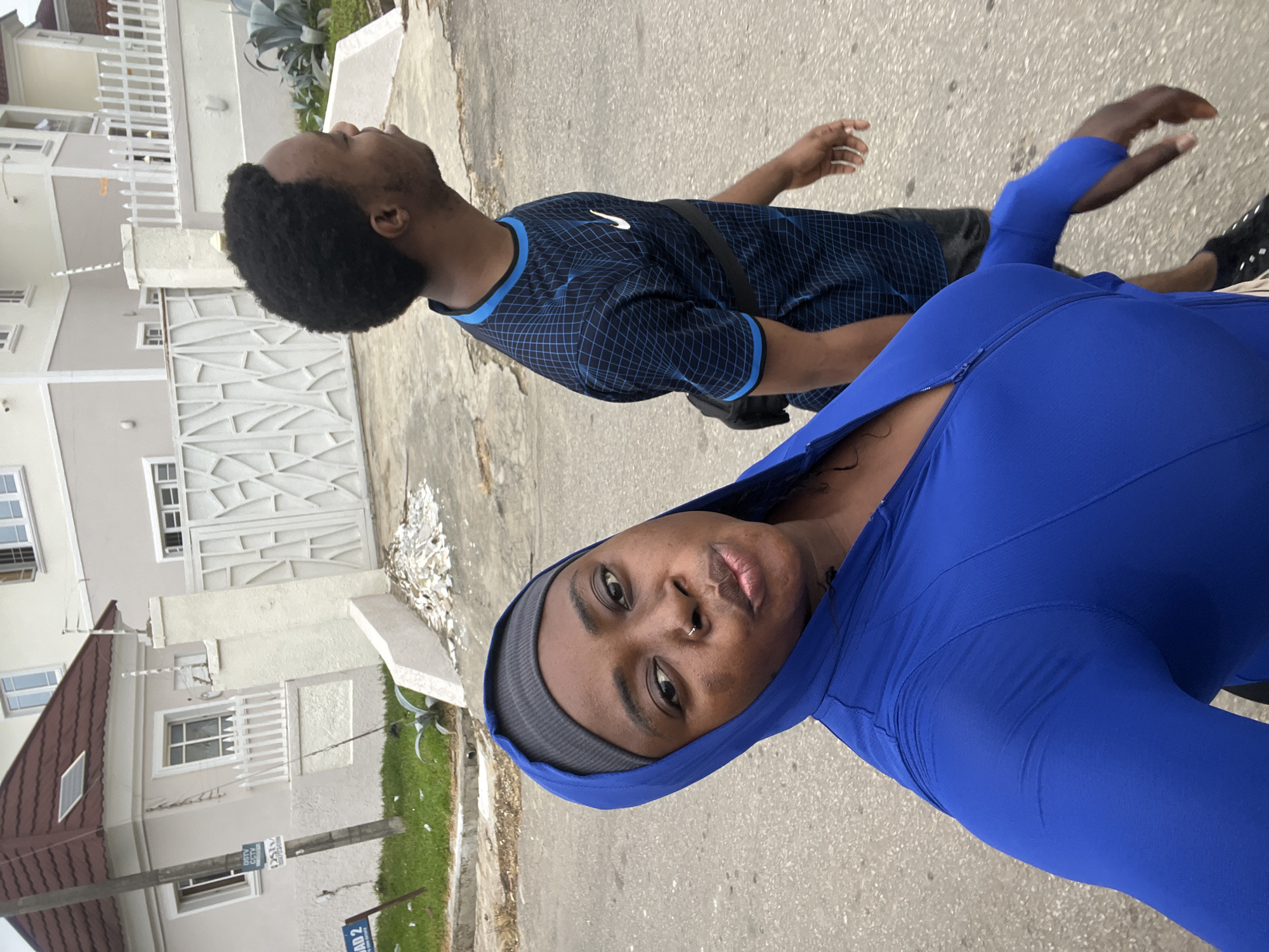 Two people, a woman in the foreground and a man behind her, are walking on a paved street in Abuja, Nigeria. The woman is wearing a blue hooded jacket and a grey headband. She is looking at the camera with her lips pursed and a nose ring. The man is wearing a blue patterned t-shirt and black shorts. He has a black fanny pack across his chest and is looking to his right. In the background, there are two multi-story buildings with white and beige facades, a white gate, and a white fence. There is also a street sign that reads "ROAD 2". The weather appears to be overcast.