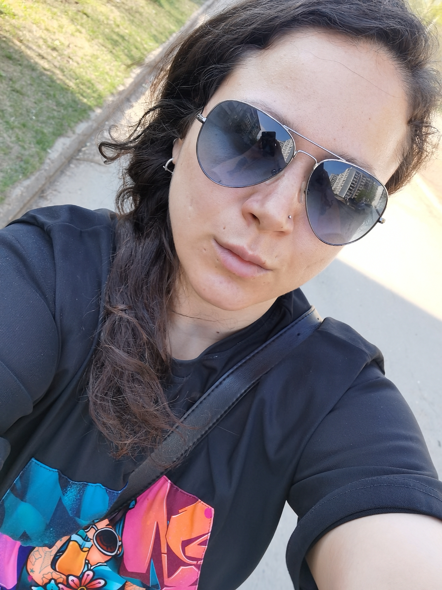 A woman wearing sunglasses and a black t-shirt with a colorful graphic is taking a selfie. She has dark hair and a nose piercing. The reflection in her sunglasses shows a multi-story building, likely an apartment complex. The photo appears to be taken outdoors on a sunny day, with a sidewalk and grass visible in the background. The location is Chaikovsky, Russia, based on the building's architectural style. A black shoulder bag is slung across her body.