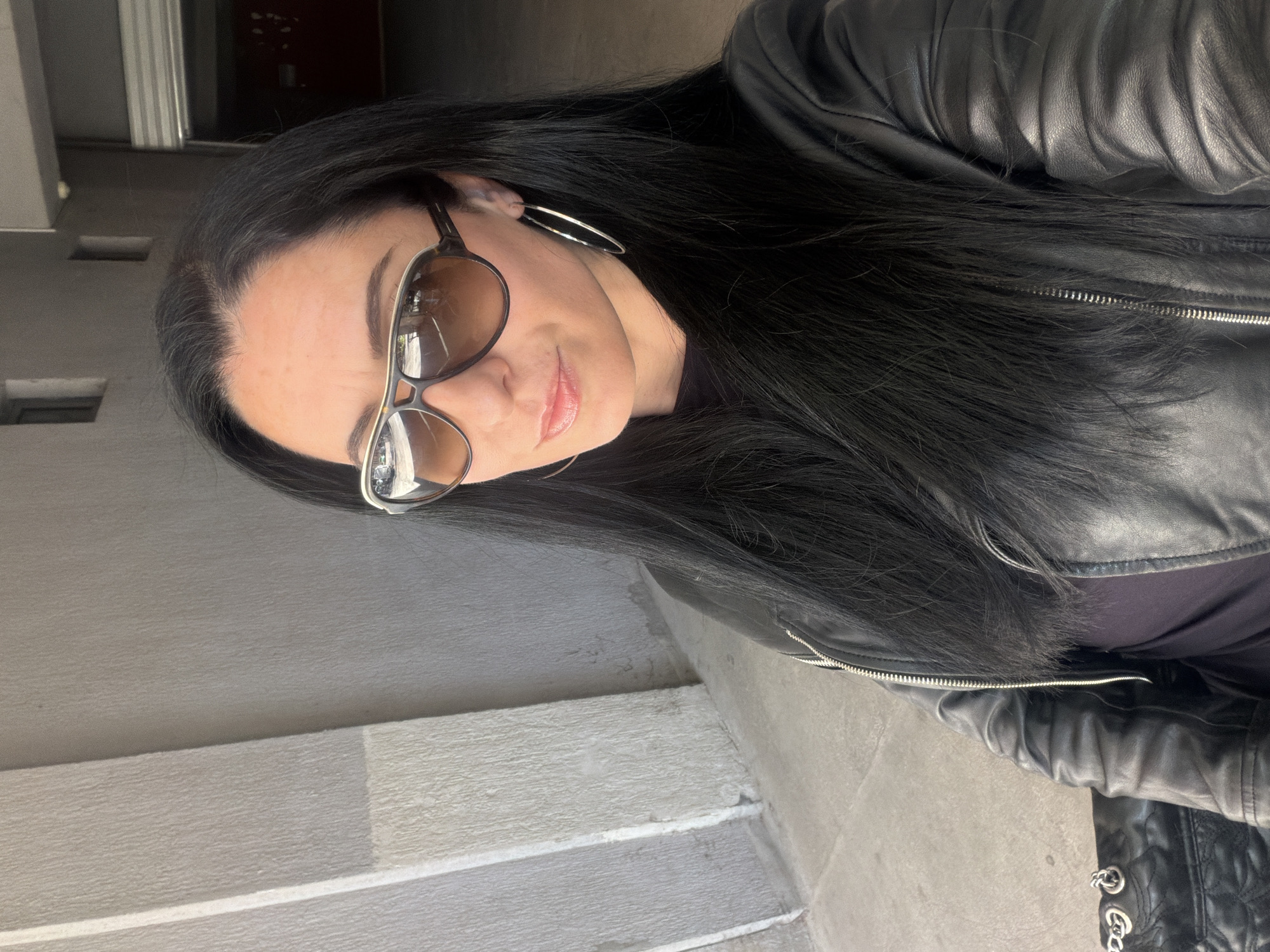 A woman is photographed in an urban environment. She has long, dark hair and is wearing sunglasses and hoop earrings. She is wearing a black leather jacket and a dark-colored top. The background shows concrete stairs and a wall. The location is the city of Pleven, Bulgaria, but there are no specific landmarks visible in the image to pinpoint the exact location.