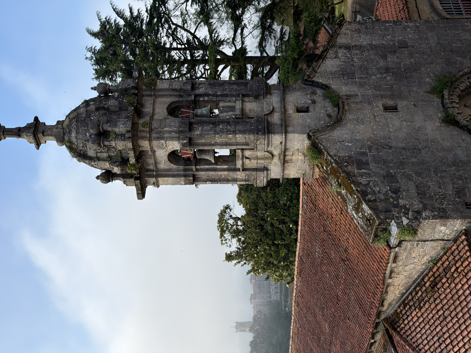 The image captures a high-angle view of a stone structure, likely a church or bell tower, with a dome-shaped roof and open arches containing bells. The structure exhibits signs of age, with moss and lichen growth on the stone. A dense tree is situated behind the tower, partially obscuring the sky. The foreground reveals a roof covered in terracotta tiles. In the background, through the fog, a building resembling a castle is visible. The location is suggestive of Santiago De Compostela, Spain, given the architectural style and landscape. There are no people visible.