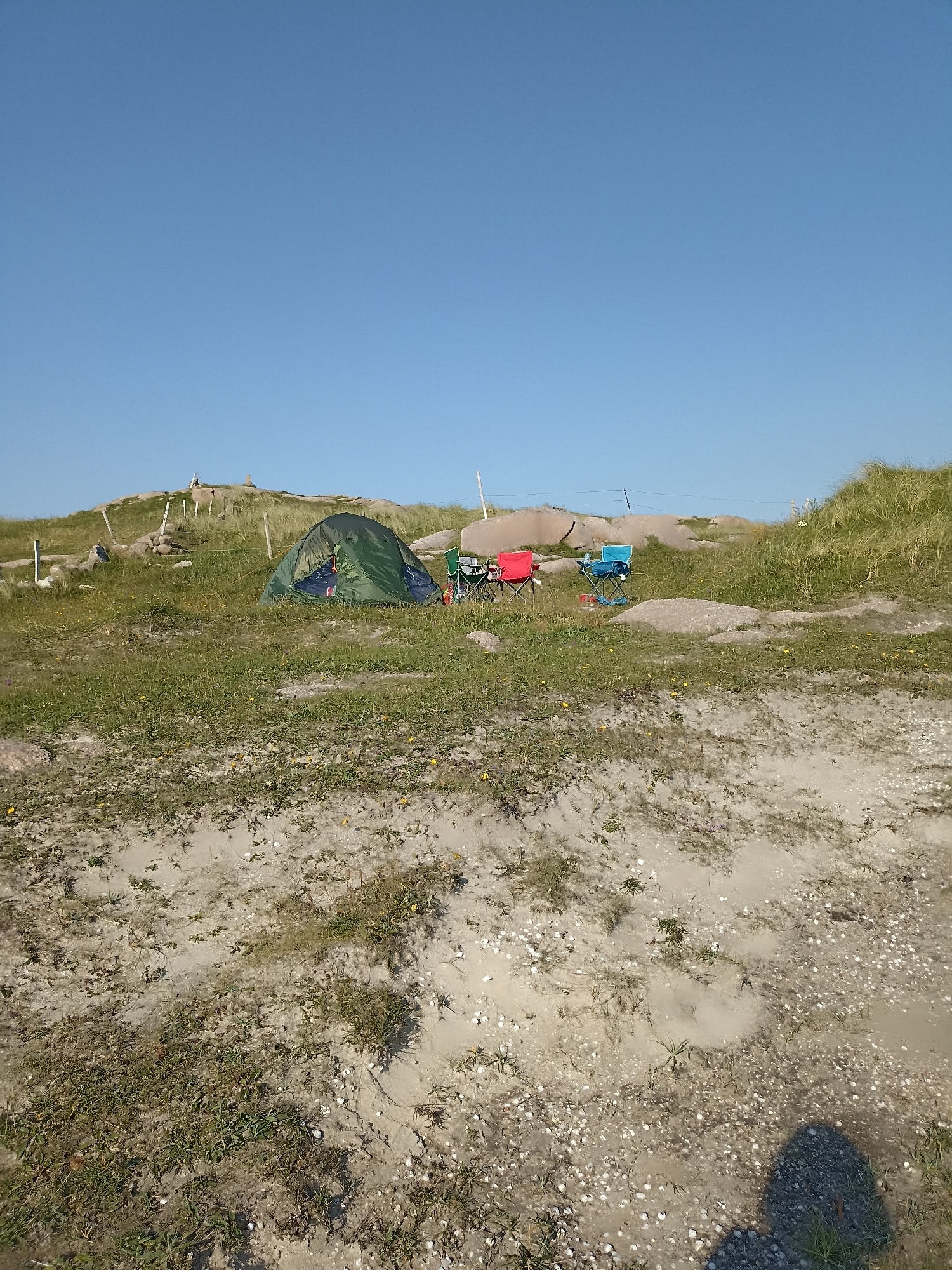 This image depicts an outdoor campsite on a grassy hillside with sandy patches, located in Carnboy, Ireland. The scene is dominated by a clear blue sky. In the middle ground, a green tent is set up, and beside it are three camping chairs: one red, one blue, and one partially visible green chair. The terrain is characterized by sparse green grass dotted with small yellow flowers, interspersed with rocks and boulders of various sizes. In the background, a gentle slope rises, covered in grass and rocks, with some faint structures and fence posts visible. The foreground consists of sandy ground with scattered shells and some vegetation. The overall impression is a tranquil, undeveloped camping spot under a bright, sunny sky.