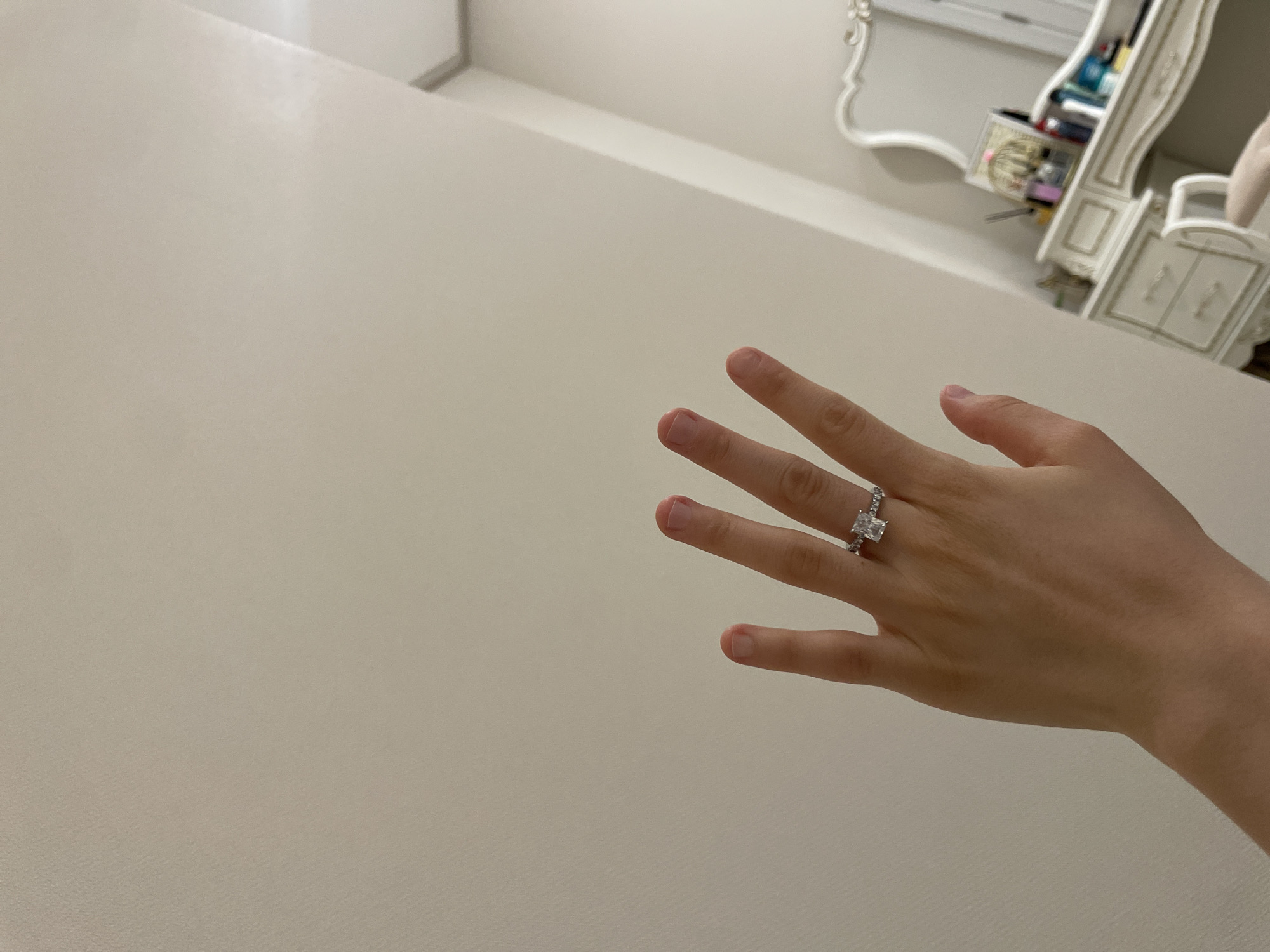 A hand with a diamond ring is positioned against a plain, off-white wall. In the background, a white vanity table with drawers and a decorative mirror is visible. Various cosmetic items are placed on the vanity. The scene appears to be an indoor setting. The location of Tashkent, Uzbekistan is not indicated by any visual elements in the image.