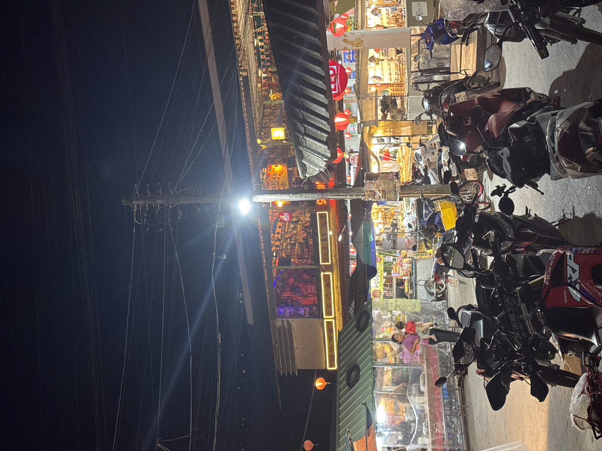 The photograph shows a street at night, likely in Thong Sala, Thailand, given the textual elements in the image. A multitude of motorcycles are parked along the right side of the frame. Above the motorcycles are numerous storefronts and shops, illuminated by bright lights and lanterns. Visible among the shops are signs in Thai, and the words "The Mexican Restaurant & Bar". Power lines are prominent on the left side of the frame, running from a tall pole into the darkness. Several individuals are visible in the shops, appearing to be browsing or working. The image is slightly blurry, possibly due to the low-light conditions and camera settings.