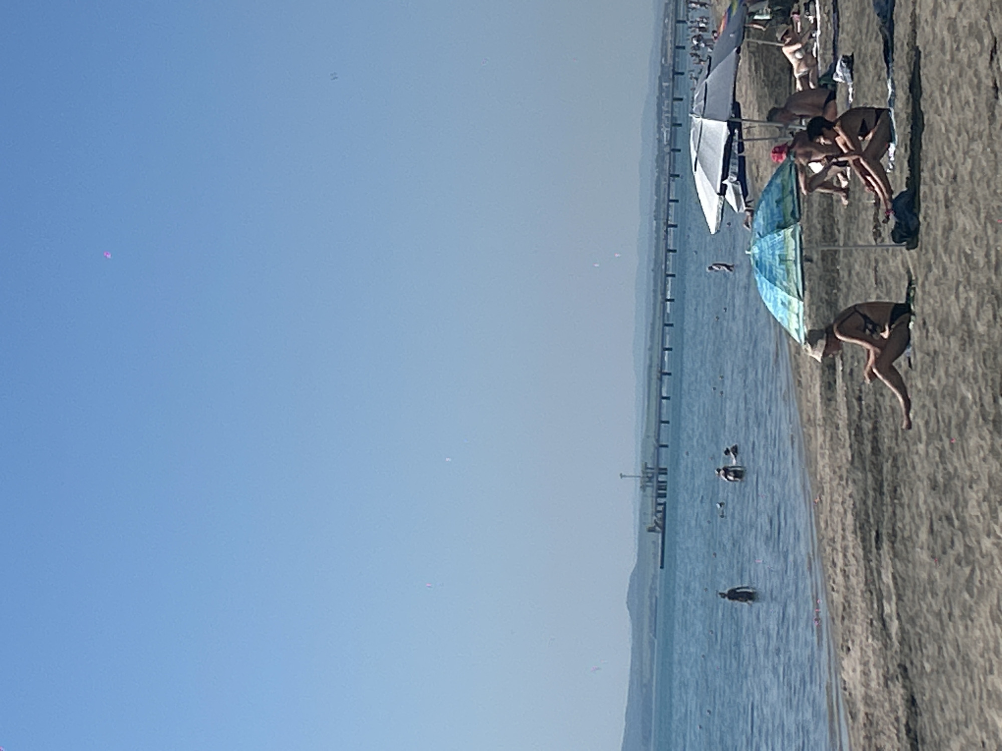 This is a photograph of a beach scene in Burgas, Bulgaria, likely during daylight hours with clear skies. In the foreground, several people are sunbathing and relaxing on the sand under beach umbrellas. Some individuals are seated, while others are lying down. The beach is sandy. In the middle ground, the sea is visible with several people wading or swimming in the water. A pier or jetty extends into the sea in the background, with structures at its end. Further in the distance, a coastline with hills or mountains is visible. The overall atmosphere suggests a typical summer day at the seaside.