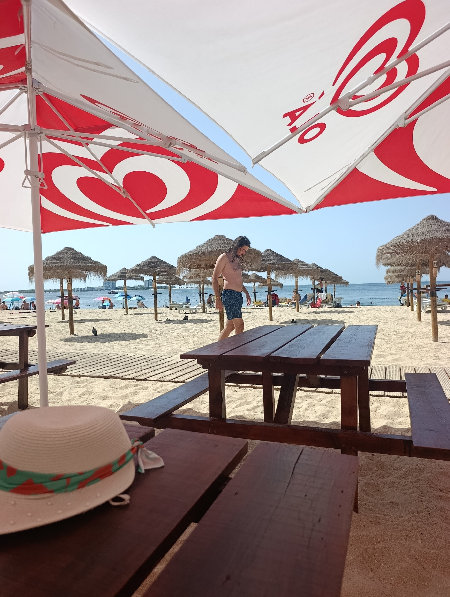 The image depicts a sunny day at Praia da Figueirinha, a beach located in Setúbal, Portugal. In the foreground, a wooden picnic table and benches are visible, with a straw hat adorned with a green and orange ribbon resting on one of the benches. Beyond the picnic area, a sandy beach stretches towards the sea. Numerous thatched umbrellas provide shade, and several people are scattered across the beach, some lounging on chairs or towels, while others are walking. A man wearing blue patterned swim trunks is walking away from the camera on the beach. The calm sea is visible in the background, with a few boats in the distance. The sky is clear and blue. The scene suggests a typical summer day at a popular beach.