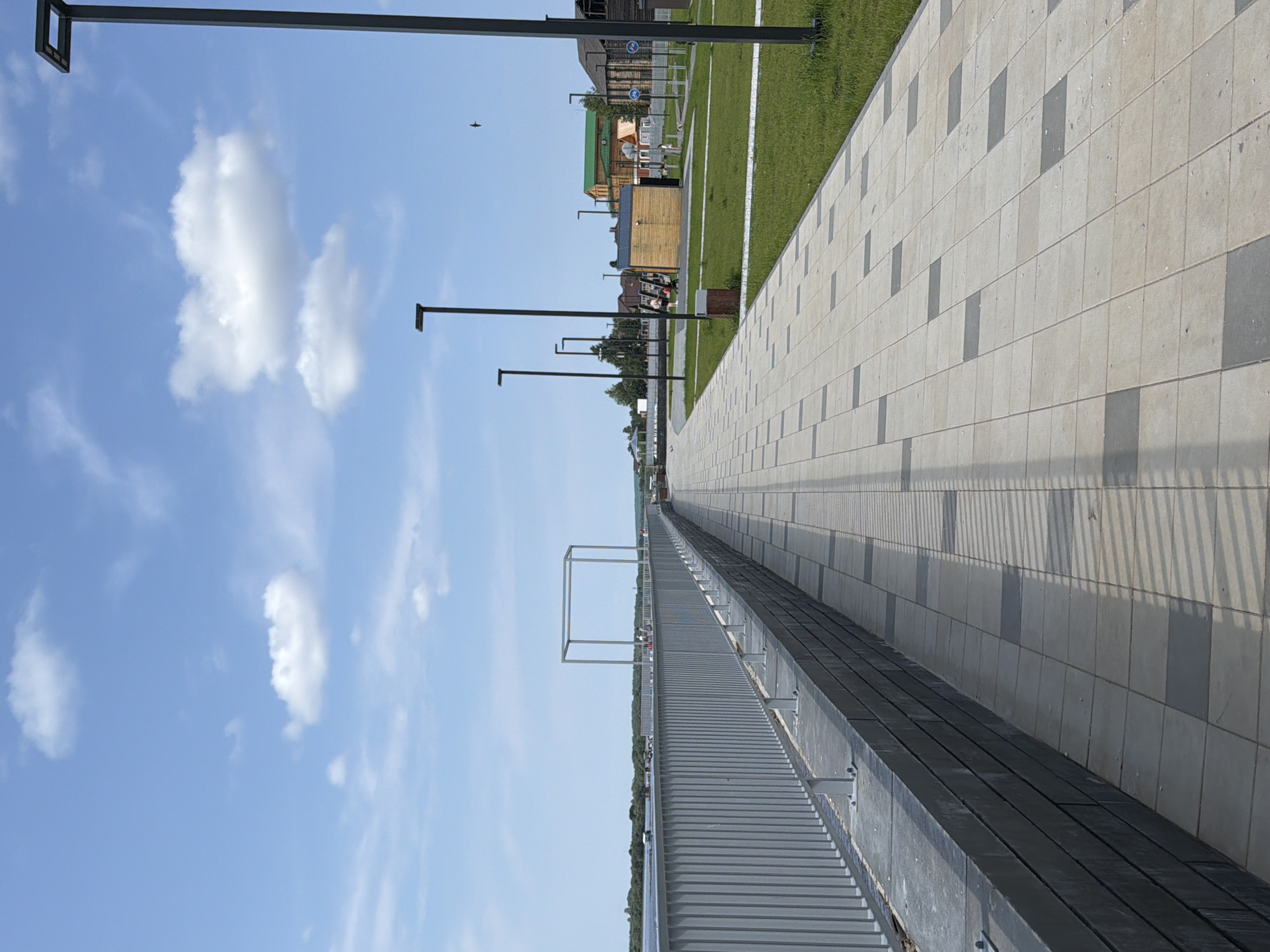 The image depicts a paved walkway bordering a body of water. The walkway is made of light-colored square tiles with darker square tiles interspersed, creating a patterned effect. A modern metal railing runs along the left side of the walkway, separating it from the water. Beyond the railing, a river or large lake is visible, with a distant treeline on the opposite bank. The sky is blue with scattered white clouds.
In the background, there is a grassy area with several modern streetlights. A small, light-brown wooden structure is also present. Further back, several buildings with pitched roofs, some with green or brown roofing, are visible. Some people are present in the scene, walking or standing in the grassy area.
The scene appears to be a public waterfront promenade. Based on the context provided, this location is in the city of Solikamsk, Russia. Specifically, this is likely a section of the embankment along the Kama River in Solikamsk, which has undergone recent development and beautification, featuring new pedestrian zones, lighting, and recreational areas.
