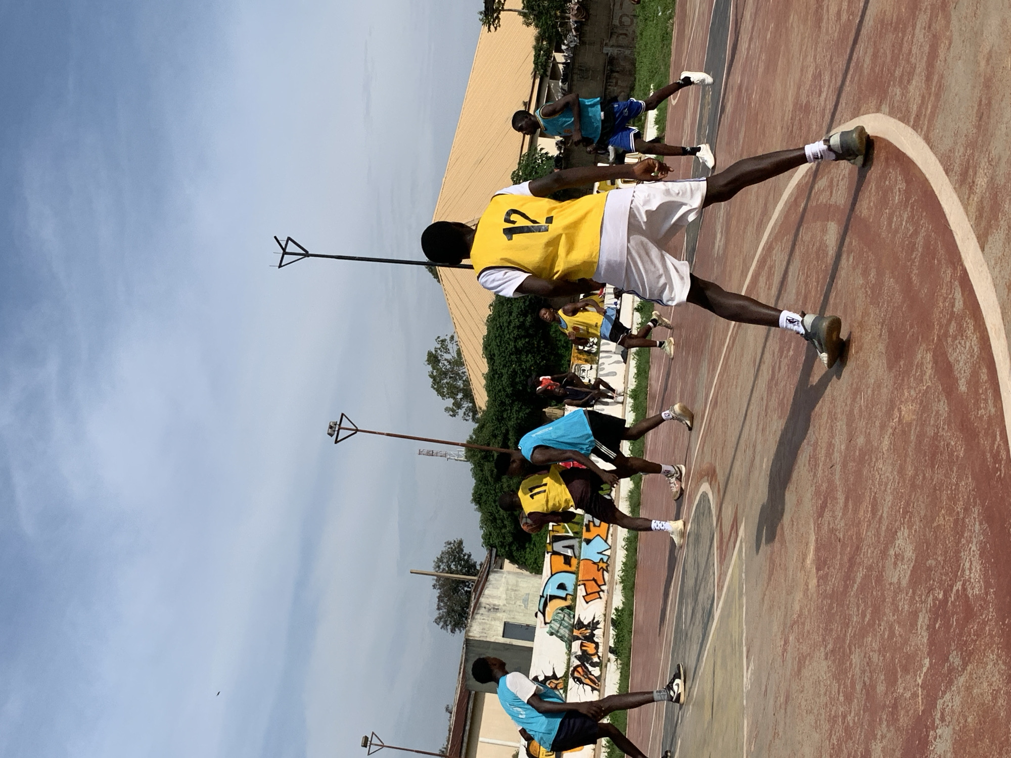 The image depicts a basketball game being played on an outdoor court. Several young men, dressed in basketball attire, are actively engaged in the game. One player in the foreground wears a yellow vest with the number "12" on the back and white shorts. Other players are visible in blue vests and shorts. The court surface appears to be asphalt with painted lines. In the background, there are buildings, trees, and a wall with graffiti. The sky is partly cloudy. The scene suggests a casual or recreational basketball match taking place within a community setting. While the image does not explicitly state the location in Jos, Nigeria, the presence of the basketball court and the style of buildings are consistent with urban environments in Nigeria.