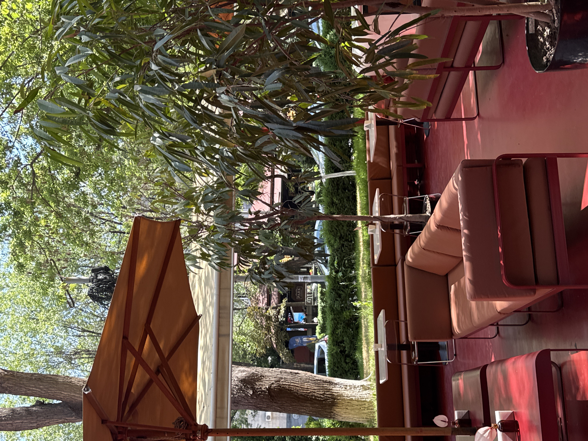 This outdoor scene captures a restaurant patio in Yerevan, Armenia. Seating comprises a series of low, brown cushioned benches with red metal frames, arranged in a row. Each bench is paired with small white side tables. A large tree provides shade to the area, and a brown sun umbrella is positioned to the left. Through the trees and foliage, a street scene is visible, featuring people, vehicles, and signs, including one that reads "PARK." The patio floor is a red hue. The perspective suggests the photograph was taken from a low angle, possibly seated at one of the tables.
