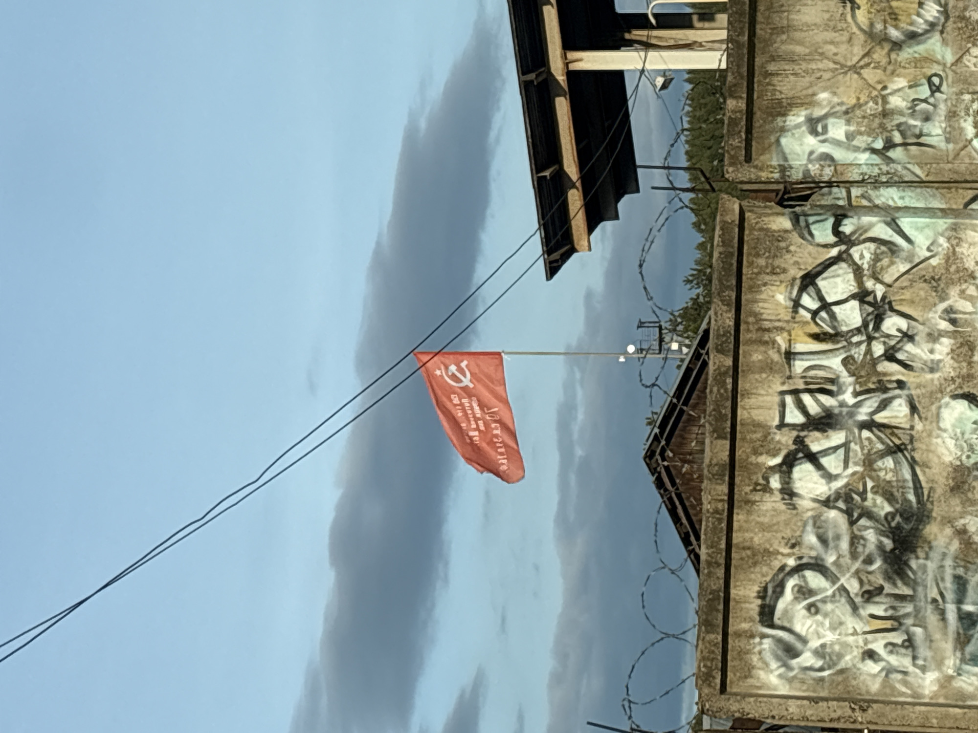 A vertical, medium shot depicts a red flag with a hammer and sickle emblem flying atop a metal pole. The flag has white writing in Russian characters. The background features a blue sky with gray clouds, utility wires, a gray building with graffiti, and barbed wire. The location is Chaikovsky, Russia.