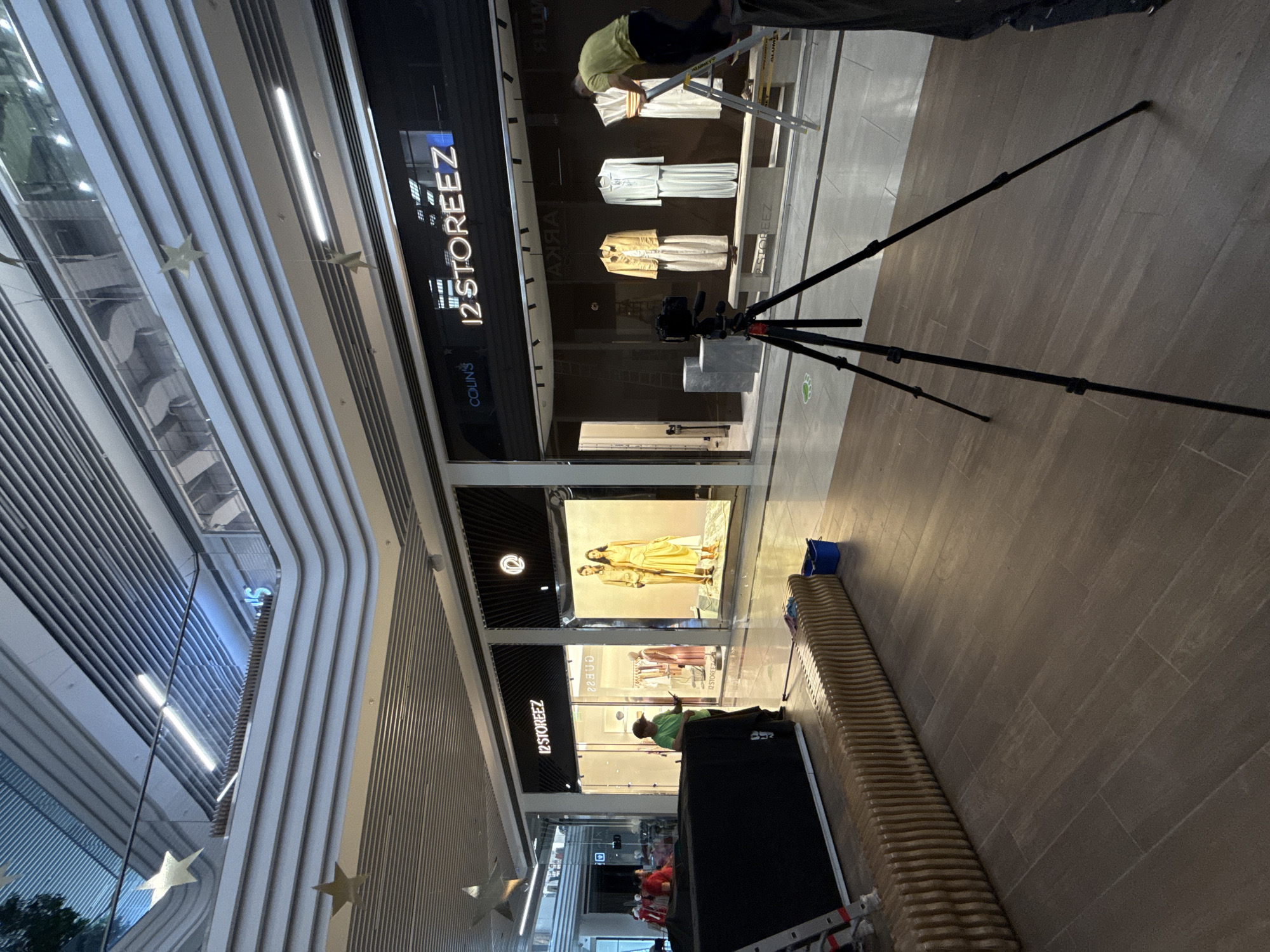 The image depicts the interior of a modern shopping mall. Visible elements include a clothing store with the name "12 STOREEZ" displayed prominently. Workers are present, possibly engaged in setting up or adjusting displays. A camera on a tripod is set up, suggesting a photoshoot is in progress. Other objects include a black equipment case, a ladder, and a blue bucket. The architecture features a multi-level design with distinctive horizontal lines and star-shaped decorations hanging from the ceiling. The mall appears well-lit and the floor is covered in brown tiles. The location is assumed to be in Perm, Russia, based on the store name "12 STOREEZ" being a Russian brand.