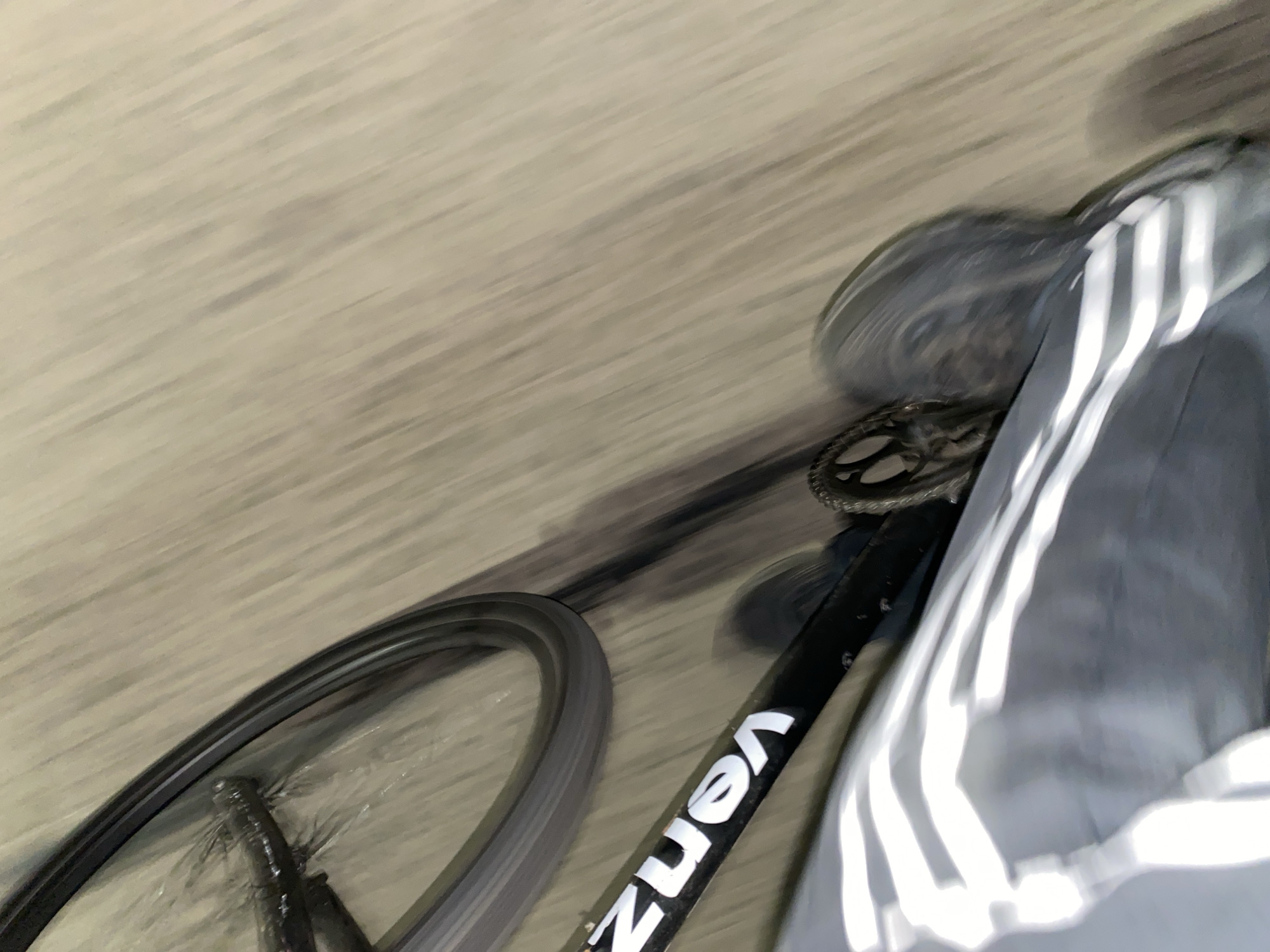 A blurry image depicts a low-angle view of a moving bicycle on a paved surface, possibly in Monte Grande, Argentina. The bicycle frame, partially visible, is black with white lettering "VENZ". A wheel spins on the left side, and the chainring is also visible. Adjacent to the bicycle is a person wearing gray pants with white stripes on the side. The surrounding environment appears to be a street or walkway with indistinct surface details due to the motion blur.