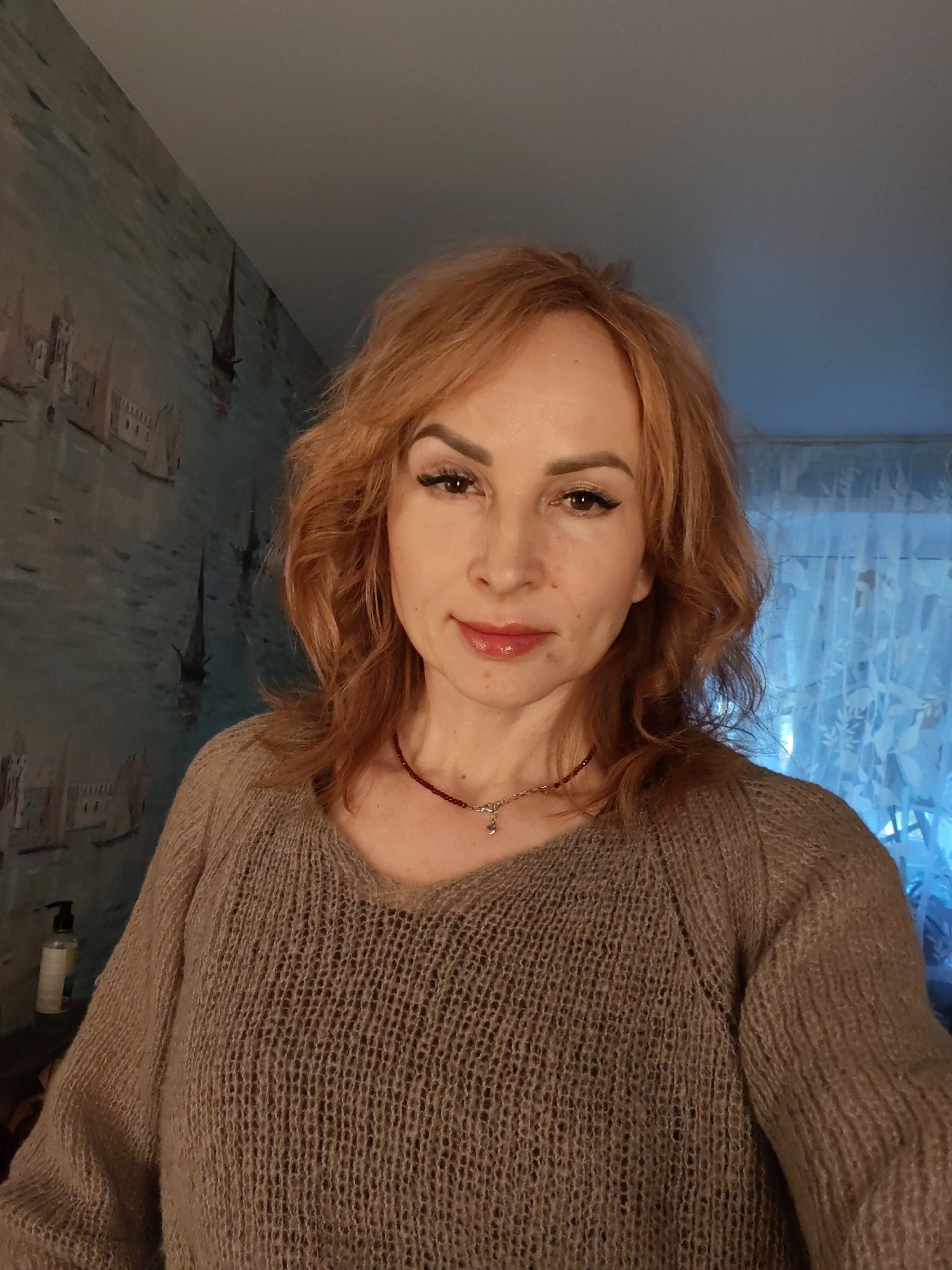 A Caucasian woman with red hair is posing for a selfie indoors. She is wearing a brown, v-neck sweater and a red beaded necklace. Her makeup includes defined eyebrows, eyeliner, and glossy lipstick. The background includes a wall with a painting of a cityscape and a window covered with sheer, floral-patterned curtains. A bottle is visible on a surface to the left of the woman. There is no explicit evidence indicating the location is Chaikovsky, Russia.