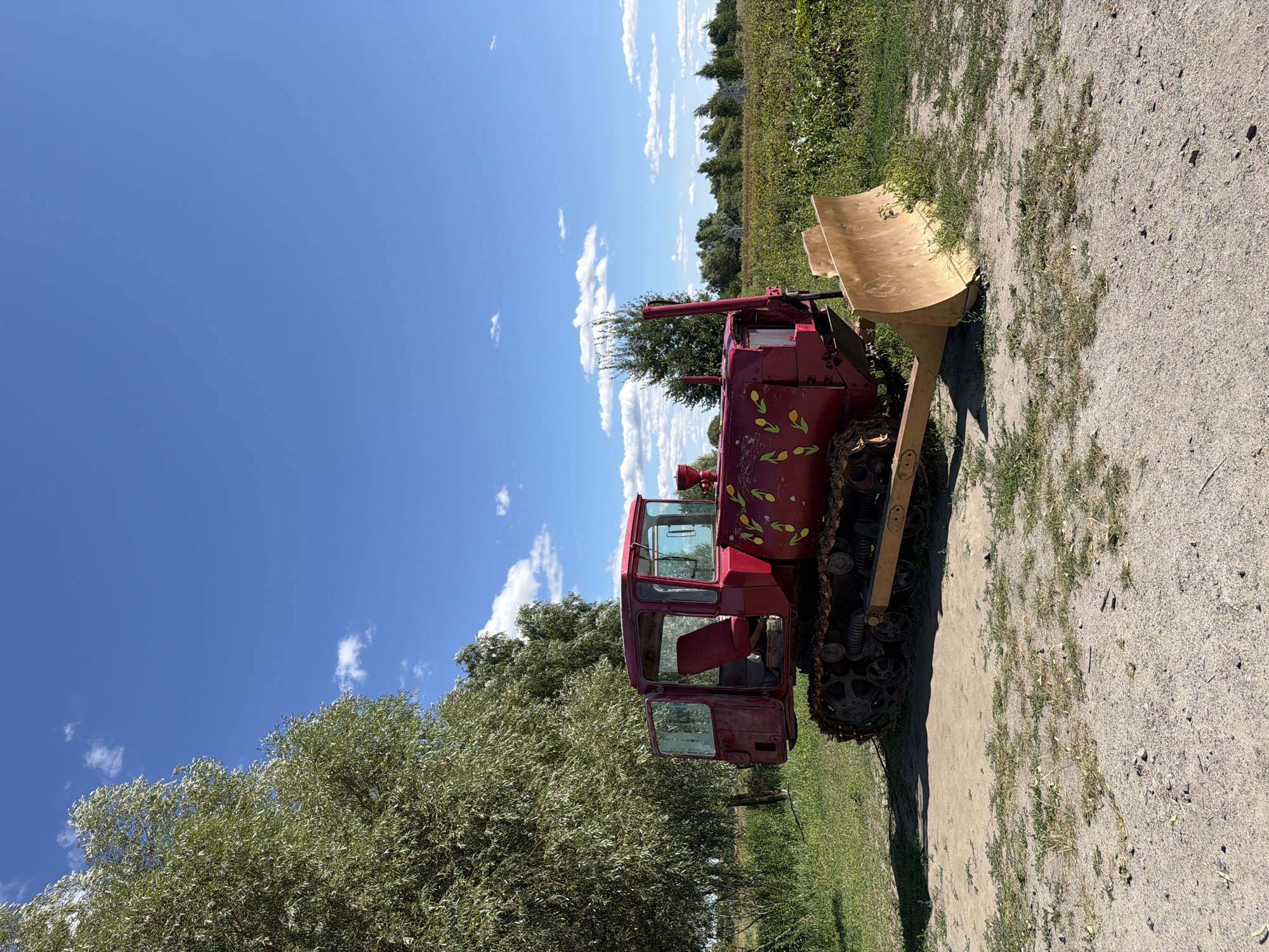 A weathered, red crawler tractor with a yellow blade sits on a dirt patch in front of a field of green plants. The tractor has a stylized pattern of green leaves and yellow shapes painted on its side. A large, leafy tree stands to the left of the tractor, with a clear blue sky dotted with white clouds above. In the background, more trees and vegetation are visible, along with two white, windmill-like structures in the distance. The scene is consistent with the rural landscape of Severynivka, Ukraine.