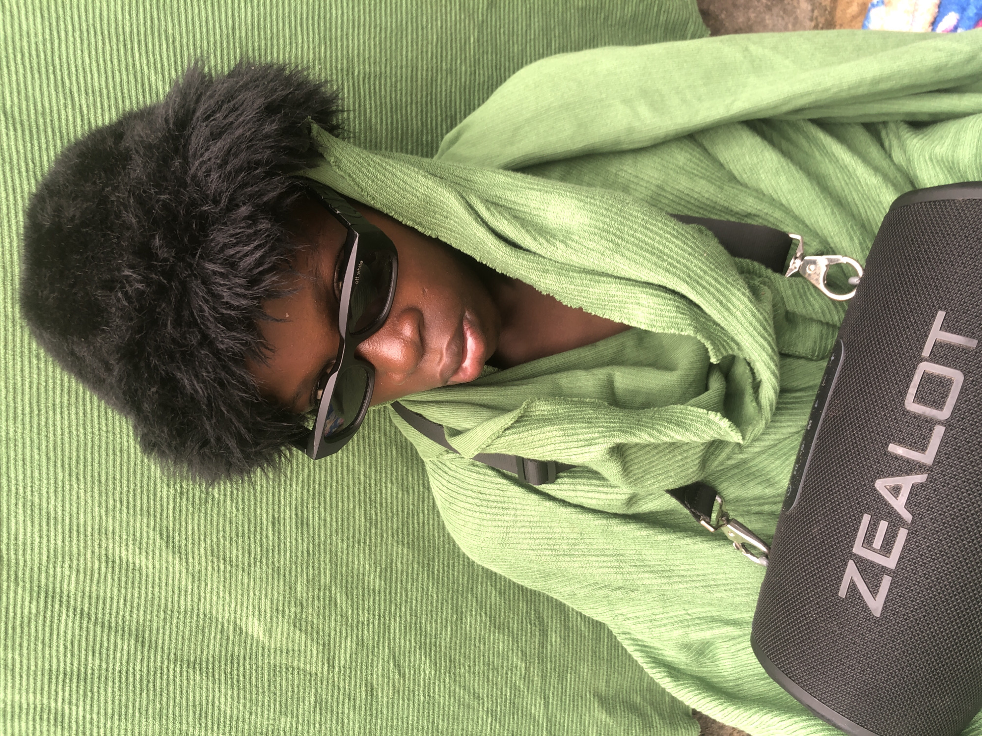 A young Black person wearing a black fuzzy hat and black sunglasses is draped in a lime green hooded garment. A black portable speaker with the word "ZEALOT" in silver letters rests in front of them, held by a black strap. The background is a textured green surface, possibly a cloth or curtain. The lighting suggests an outdoor or brightly lit indoor setting. No specific landmarks or elements identifying Ado, Nigeria, are visible in the image.