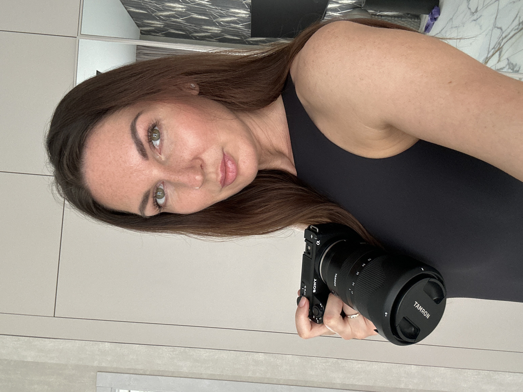 The image, rotated 90 degrees counter-clockwise, displays a woman taking a mirror selfie indoors. The woman, appearing to be in her 20s or 30s, has long, straight, dark brown hair that falls over her shoulder. She has fair skin, noticeable freckles across her nose and cheeks, and striking light green or hazel eyes. Her eyebrows are well-defined, and she wears subtle makeup, including what appears to be lip gloss or a light lipstick. She looks directly at the camera with a calm expression, featuring a slight, subtle smile.
She is wearing a simple black sleeveless top or dress, visible over her left shoulder and arm. Her right hand, with neatly manicured nails and a silver-colored ring on her ring finger, holds a black mirrorless camera. The camera body clearly shows the "SONY" brand name, and an "α" (Alpha) logo is visible on the hot shoe mount. Attached to the camera is a black "TAMRON" zoom lens, which has focal length markings at "17", "24", "35", "50", and "70". The lens cap, also black, prominently features the "TAMRON" logo.
The background consists of light-colored, possibly beige or off-white, wall paneling with visible seams. Above her head (to the left in the rotated image), a darker, patterned surface, possibly a decorative wall or a reflection in a mirror, adds a textural element to the setting. The general impression is of a modern, well-lit interior, likely a home or apartment in Chaikovsky, Russia. The woman's activity suggests an interest in photography or showcasing her camera.