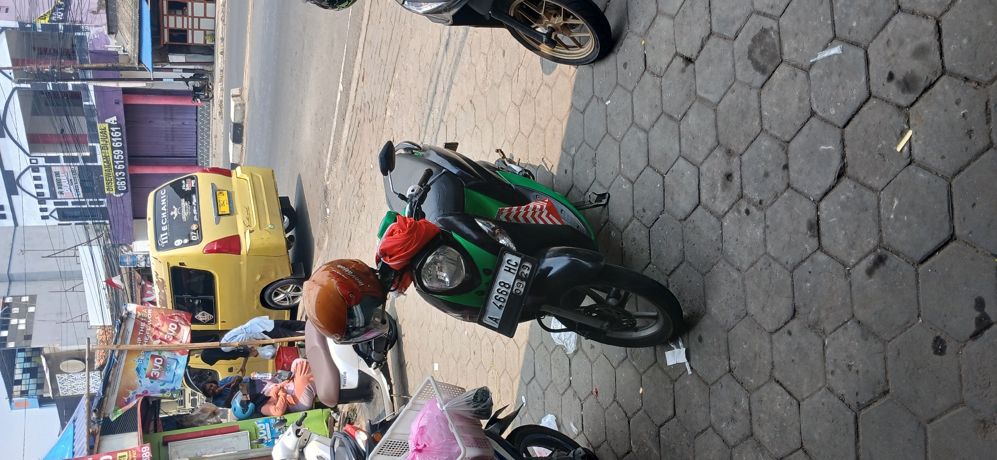 In Baros, Indonesia, a busy street scene unfolds. In the foreground, a green and black scooter is parked on a gray hexagonal-tiled sidewalk. A brown helmet with red and white accents sits on the handlebars, adorned with a red cloth. The scooter's license plate reads "A 4668 HC". To the left, a white scooter with a brown seat is parked, and a white basket filled with pink and clear plastic bags rests in front of it. A person wearing a light orange top and a blue helmet sits on the white scooter, facing away from the camera, with another person partially visible behind them.
In the background, a bright yellow van is parked on the street, displaying a "MECHANIC" decal and a license plate that appears to be "A 1842 DN". A sign on the van reads "DISewakan" (For Rent). Behind the van, buildings line the street. One building has a purple facade with large closed metal doors and a sign that says "0813 6159 6161 A". Another building to the left features a sign with an image of a drink and text that is partially obscured. The overall environment suggests a commercial area with vehicles and pedestrians.