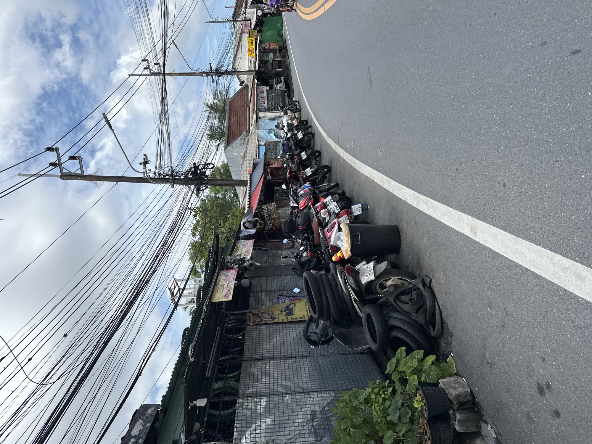 A row of parked motorcycles lines the left side of a street in an urban setting. In the foreground, a dark-colored motorcycle is laden with stacked tires. To the left of the motorcycle, several potted plants are visible. A black trash bin stands next to the motorcycle. The street is paved and features a white line marking the edge. Overhead, a complex network of electrical wires crisscrosses the sky, supported by utility poles. Buildings with corrugated metal roofs are visible along the street, some displaying signage. The sky is partly cloudy.