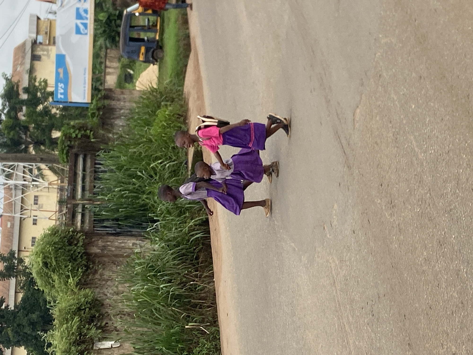 The image depicts three young children in school uniforms crossing a paved road in Nkpor, Nigeria. The two older children are wearing matching purple dresses with white collars and short sleeves, and one carries a black backpack. The youngest child, in the middle, wears a similar purple dress. The child on the right wears a bright pink shirt and a purple skirt with a patterned hem. They are walking from left to right across the frame, with the two older children holding hands. The road surface is asphalt with some lighter patches and tire marks. In the background, there is a stone wall with dense green foliage growing over it. Beyond the wall, a building with a red-tiled roof and a white metal structure are visible. A blue and yellow "TVS" advertisement banner is also present. To the right of the advertisement, a small blue and yellow auto-rickshaw is parked on the side of the road. Further to the right, another person in a red and yellow top is visible, partially obscured. The scene appears to be in a somewhat rural or semi-urban part of Nkpor, with elements of both natural vegetation and man-made structures.