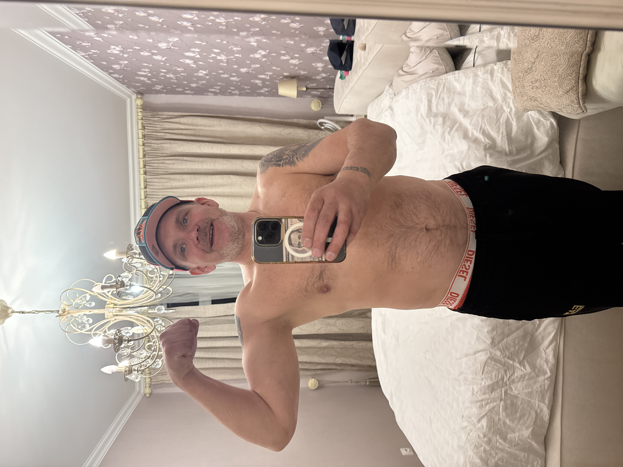 A man, appearing to be in his 30s or 40s, is captured in a mirror selfie, shirtless and smiling at the camera while flexing his left arm. He has a short beard or stubble, blue eyes, and is wearing a light-colored baseball cap with pink and blue accents. Tattoos are visible on his left shoulder and forearm. He holds a gold-framed smartphone in his right hand, equipped with a phone case that features a portrait, possibly resembling a currency note. He is wearing black shorts or underwear, with the "DIESEL" logo prominently displayed in red and white on the waistband, appearing reversed due to the mirror reflection.
The setting is an indoor room, likely a bedroom in Saint Petersburg, Russia, as reflected in the mirror. An ornate, gold-colored chandelier with multiple lights and crystal decorations hangs from the ceiling. The walls feature a light-colored wallpaper with a subtle floral or leaf pattern above plainer, lighter sections. Heavy, light brown curtains are drawn. To the man's right, a large bed with white bedding and several pillows (white, grey, and one with a beige pattern) is visible. A nightstand with a lamp and some dark, folded clothes are also reflected in the background. The lighting suggests an indoor setting, likely evening or artificial illumination. The man's expression conveys a sense of happiness or confidence.