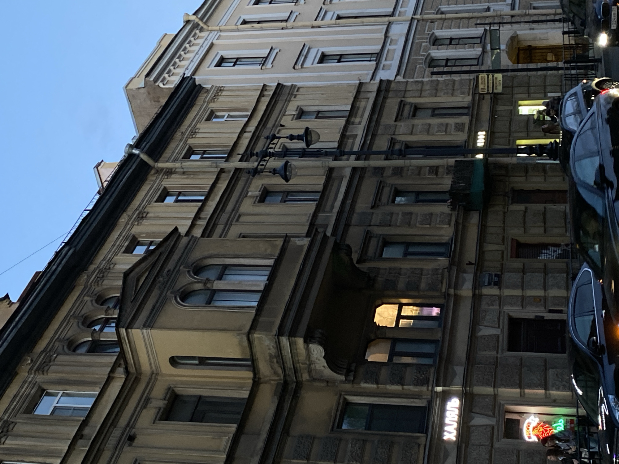 A low-angle shot captures a street scene in Saint Petersburg, Russia, primarily featuring the facade of a historic building. The building is characterized by its ornate architectural details, including decorative windows, balconies, and textured stonework. A street lamp extends upwards, partially obscuring the building.
Parked vehicles, including a black car, are visible on the right side of the frame, reflecting light from the street. A neon sign, possibly indicating a business, is present on the building's ground floor. The sky is a pale blue, suggesting either early morning or late evening.
The overall environment is urban, with a focus on the architectural characteristics of the city. No people are distinctly visible, although there could be silhouettes or reflections in the vehicles. The image gives a sense of the city's historical architecture and street life.