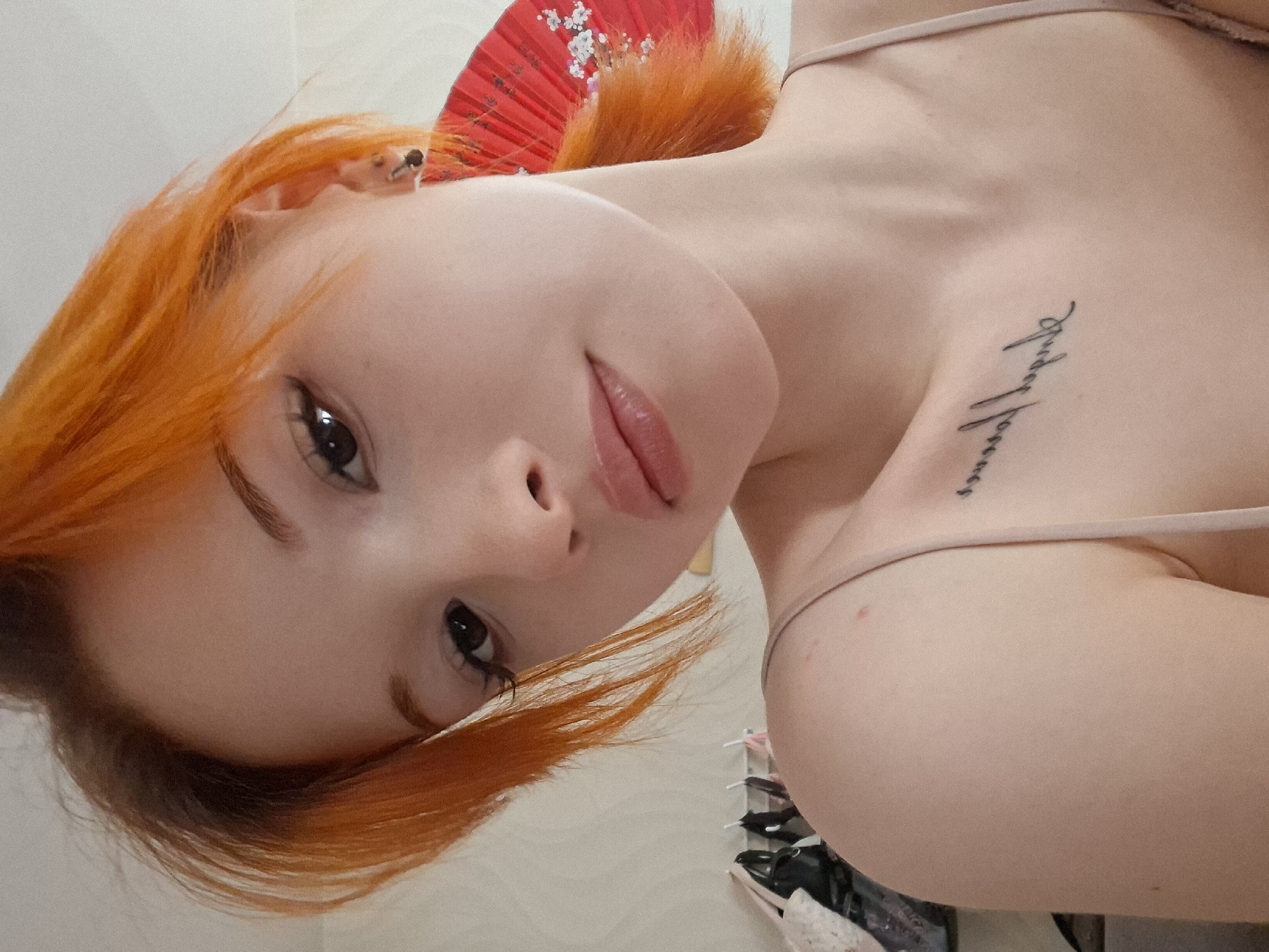 A fair-skinned person with orange hair is positioned in front of a light-colored wall. They are lying on their side, facing the camera with their head tilted. They have a tattoo on their upper chest. Behind their head, a red fan with a floral design is visible. Clothing items are partially visible in the lower right corner of the image. The location of Chaikovsky, Russia is not verifiable from the image.