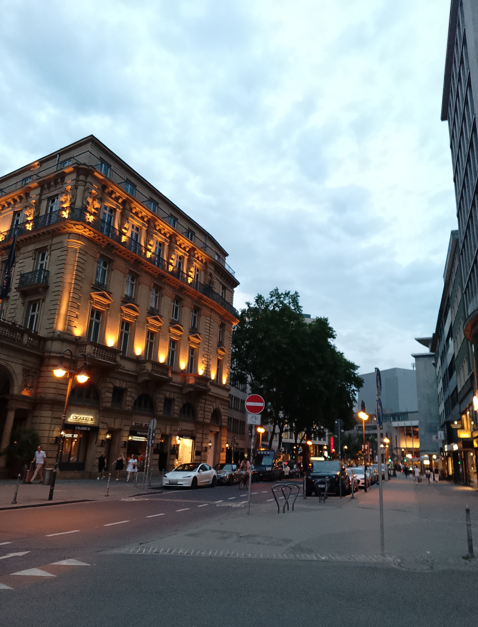 An eye-level, full shot captures a street scene in the city of Unknown at dusk. The perspective is from the street, looking towards a large, ornate building on the left and a modern building on the right. The sky is overcast with a mix of grey and white clouds.
The building on the left is a grand, multi-story structure with classical architectural elements. Its facade is illuminated by warm, yellow lights, highlighting intricate carvings and balconies. Multiple windows are visible, some appearing lit from within. A street lamp with two orange lights stands in front of this building. Below, several shop signs are visible, including one that reads "Cigarrum." People are walking on the sidewalk in front of this building. A white Tesla Model 3 is parked on the street in front of the building on the right.
The building on the right is a tall, contemporary structure with a dark facade and numerous windows arranged in a grid pattern. More shops and businesses are visible along the ground floor of this building, with illuminated signs and windows. The street is paved with asphalt and marked with white lines. Several cars are parked along the street, and traffic signs, including a red and white circular sign with a horizontal bar and a blue circular sign with a white arrow, are present. A large, dark green tree stands between the two buildings, partially obscuring the view of structures behind it. The overall atmosphere suggests a busy urban environment transitioning from day to night.