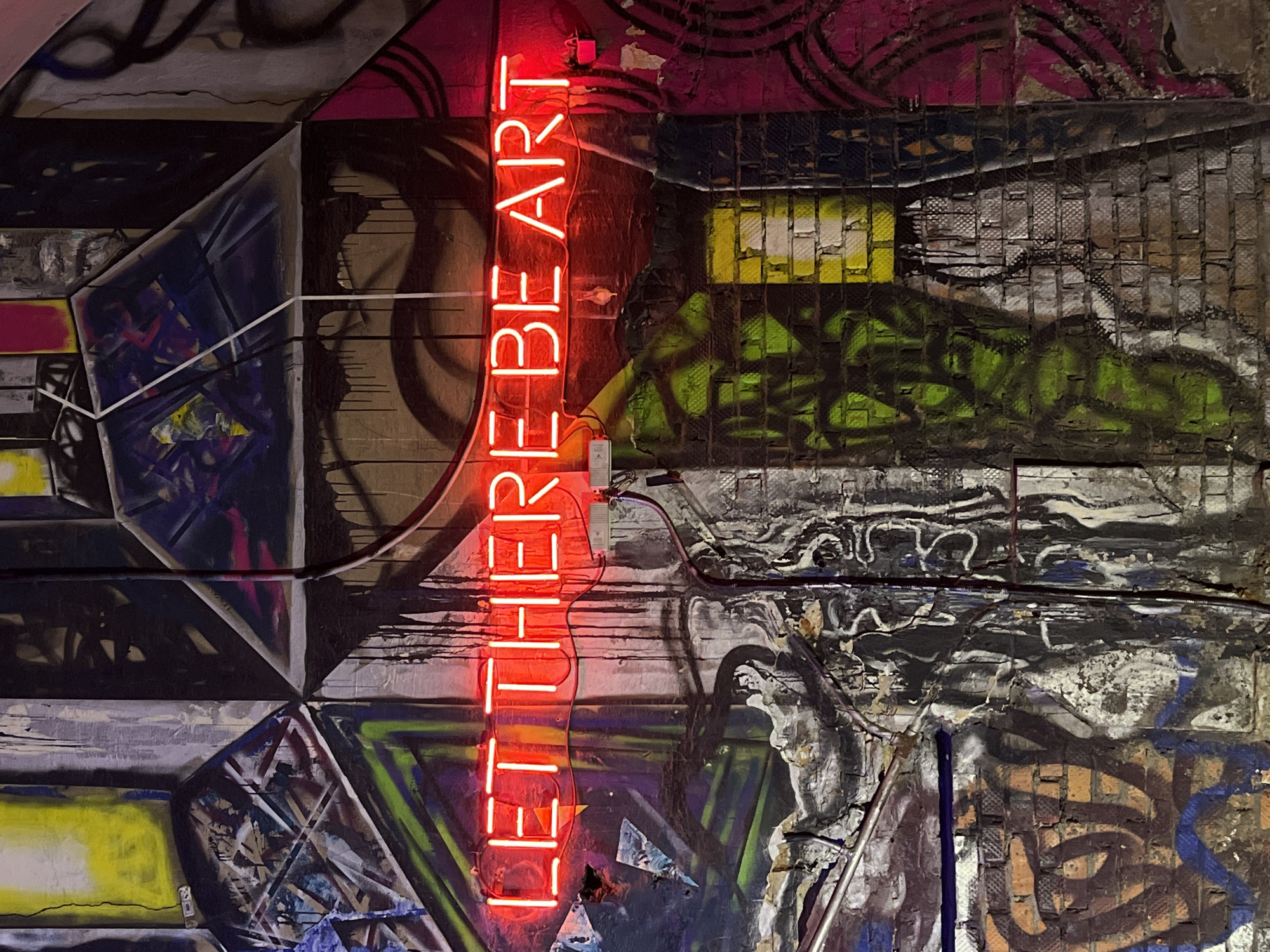 The image depicts a wall with graffiti and a neon sign that reads "LET THERE BE ART". The wall has various layers of graffiti in different colors, including black, green, pink, yellow, and blue. The graffiti is a mix of geometric shapes, abstract designs, and possibly some figurative elements. The wall appears to be made of individual sections or panels. The neon sign is vertically oriented and stands out prominently against the background. Wires are connected to the sign. The overall environment is gritty and urban. The image could be in Saint Petersburg, Russia, as the street art scene is prevalent in the city. The lighting suggests an indoor environment, possibly a gallery, bar, or abandoned building.