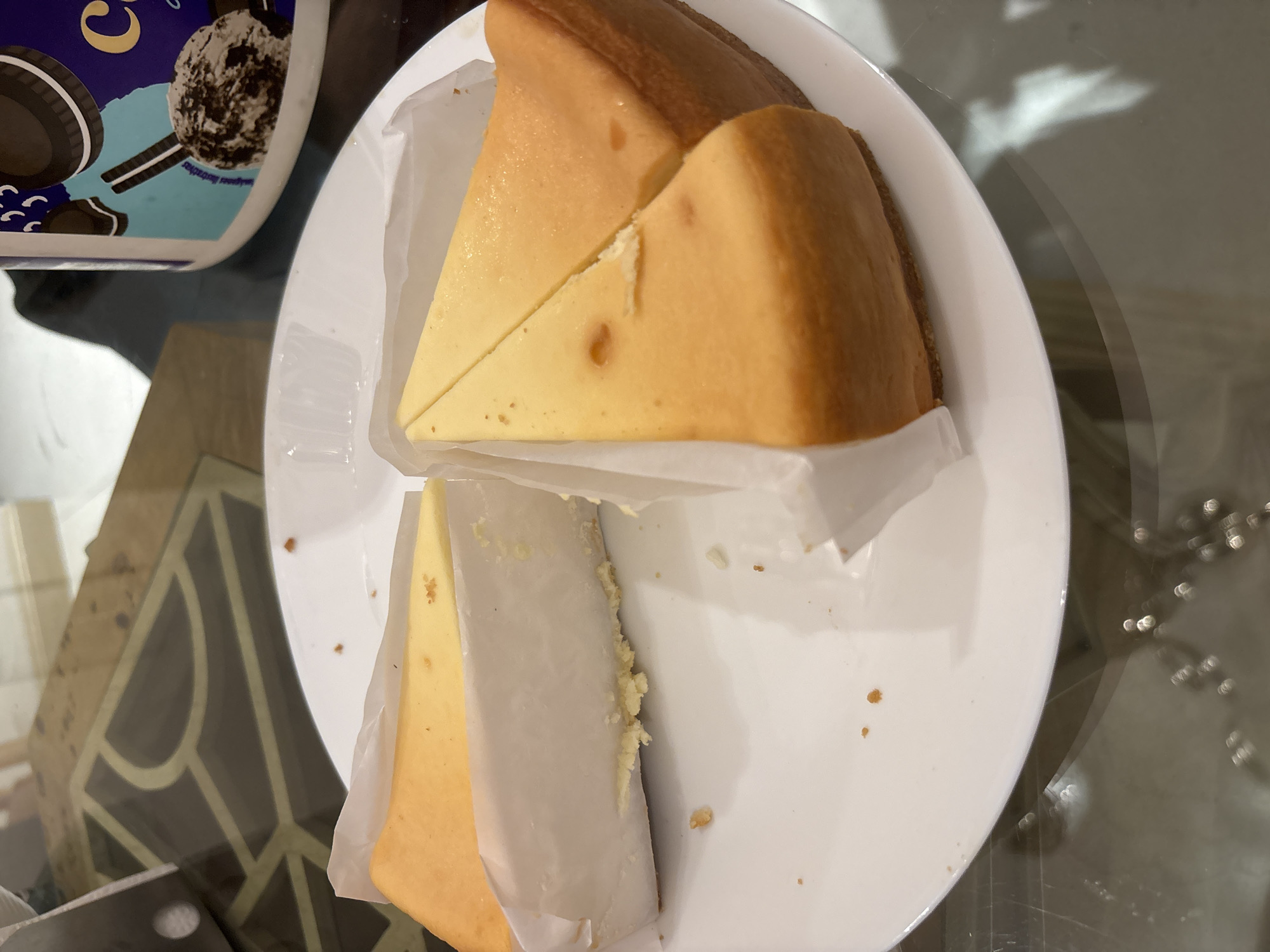 The image displays four slices of cheesecake on a white circular plate. Each cheesecake slice is partially wrapped in white parchment paper, revealing a golden-brown baked top and a lighter yellow interior. Three slices are positioned together, forming a segment of a larger cake, with the fourth slice placed slightly apart. Scattered crumbs are visible on the white plate. The plate rests on a reflective glass table, through which a dark, geometric pattern is visible beneath the surface. In the upper right background, a white plastic tub with a blue label is partially visible, featuring Oreo cookie branding and an image of cookies and cream ice cream. The scene is indoors. No individuals are present. No specific geographic identifiers for Mexico City, Mexico, are discernible within the frame.