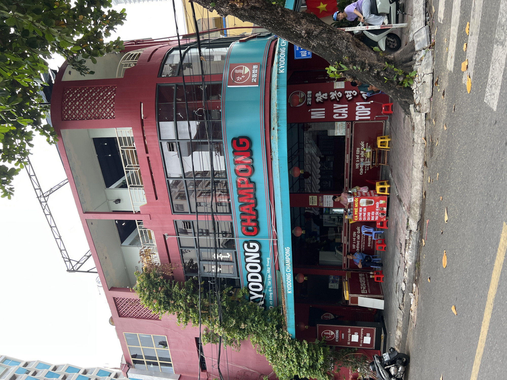 A red, multi-story building with a teal and red sign for "KYODONG CHAMPONG" stands on a street in Nha Trang, Vietnam. The building has large windows and a curved facade. Green ivy climbs the side of the building. Several people are visible sitting on small red and yellow stools outside the establishment. A motorcycle is parked on the left side of the image. A woman is sitting on a scooter to the right of the building, and a yellow and red Vietnamese flag is visible on the right. The street in front of the building has a crosswalk and fallen leaves.