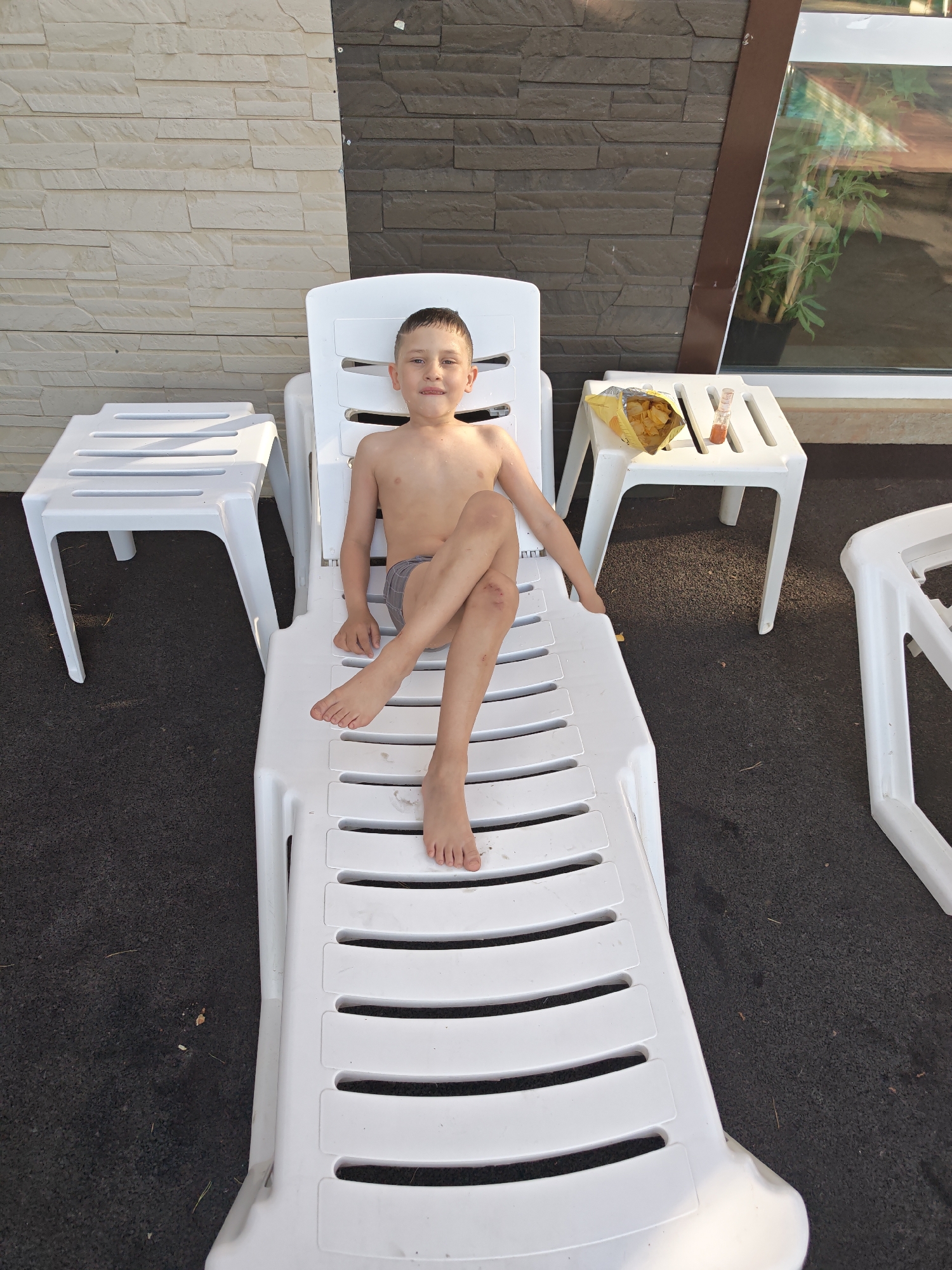 A shirtless young boy lies on a white plastic deck chair. He is wearing gray shorts. His legs are crossed at the knee. He appears to have a scrape or injury on his left knee. Next to the chair, on a small white table, is an open bag of potato chips and a small glass bottle with a brown liquid. Another similar chair is visible to the left of the deck chair. The background shows a wall with two different types of stone textures and a window reflecting greenery. The ground is dark and appears to be pavement. The image was taken in Chaikovsky, Russia.