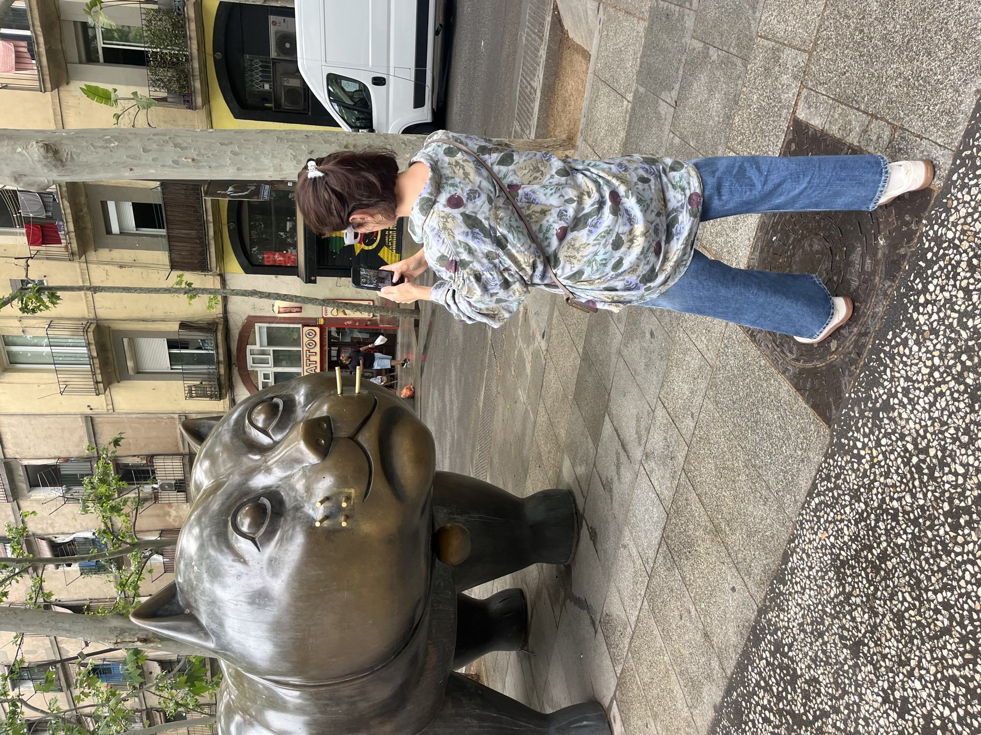 A middle-aged woman stands on a sidewalk in Barcelona, Spain, taking a picture with her smartphone of the El Gato del Raval cat statue. The woman, with brown hair pulled back with a white clip, wears a floral print blouse, blue jeans, and white sneakers with brown soles. She holds the phone in both hands, her focus on capturing the large, bronze-colored cat statue.
The statue, a prominent landmark in the Raval neighborhood, is the primary subject. The surrounding environment includes a paved sidewalk, a city street with a white van, and buildings with apartments and businesses, including a tattoo parlor with the word "TATTOO" visible on its storefront sign. A tree with green foliage grows nearby.
