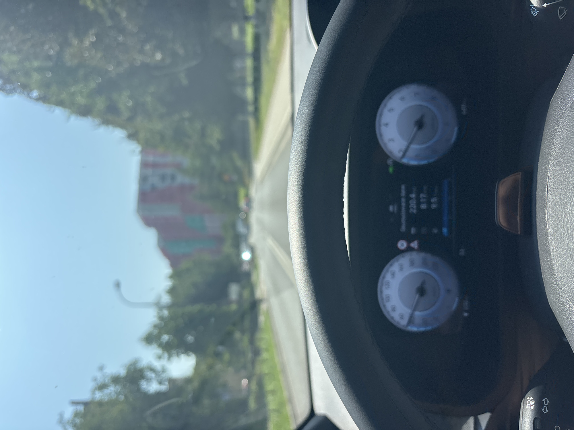 This is a first-person perspective photo taken from inside a vehicle, looking out through the windshield at a road in Krakow, Poland. The foreground is dominated by the car's steering wheel and digital dashboard display. The dashboard shows a digital speedometer reading around 95 km/h and other vehicle information. Outside, the road stretches ahead, bordered by trees on both sides. In the distance, buildings with red and green facades are visible. The sky is clear and blue, indicating a sunny day. The overall impression is that of driving through a suburban or semi-urban area of the city.