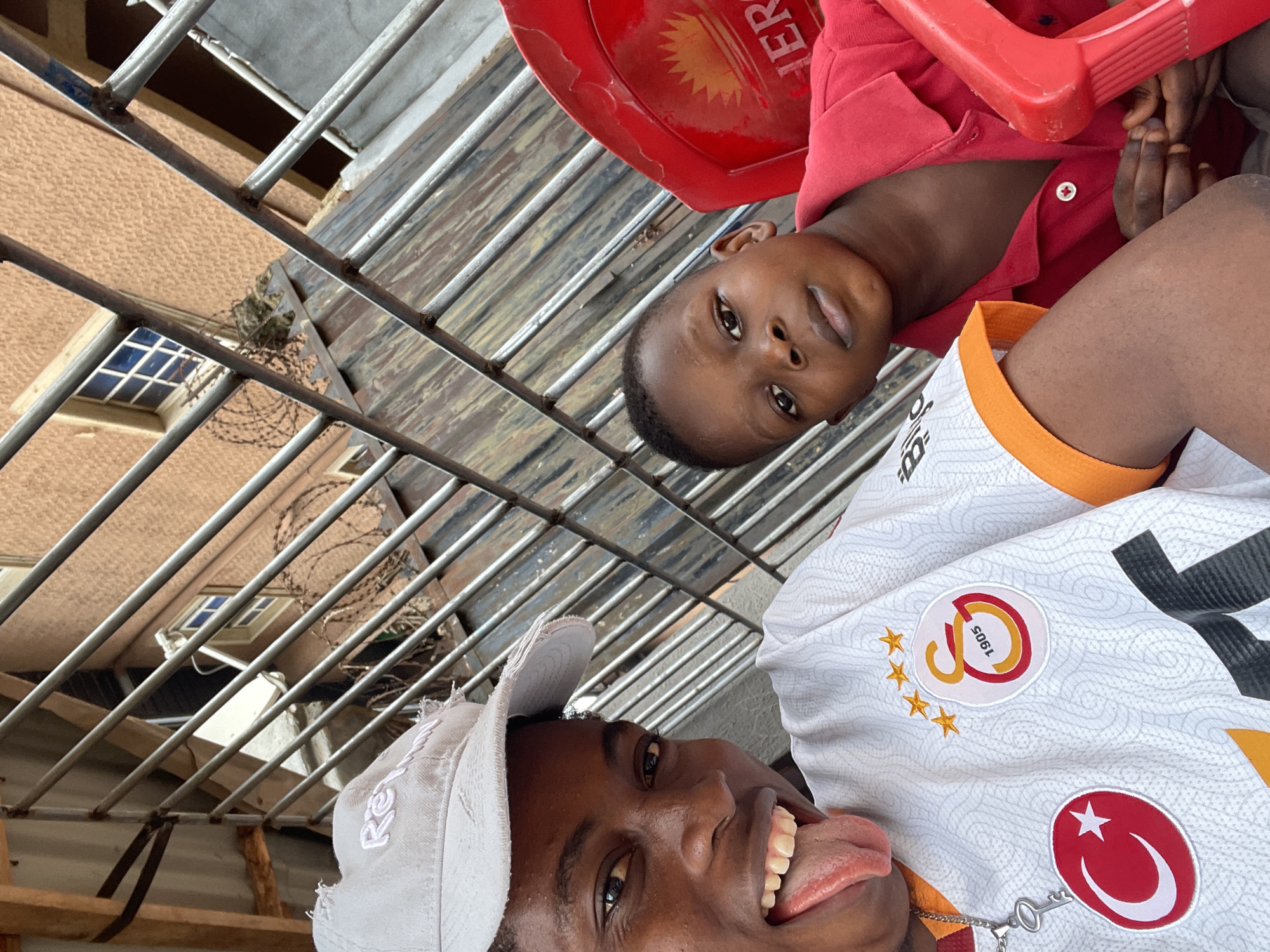 Two individuals are in the frame. The person in the foreground is wearing a light grey cap and a white soccer jersey, with the Galatasaray logo and Turkish flag visible. This person has their tongue extended. The second person is a child wearing a red shirt, seated in a red plastic chair. The background features a building with barred windows, barbed wire along the fence. The location is suggested to be Onitsha, Nigeria.