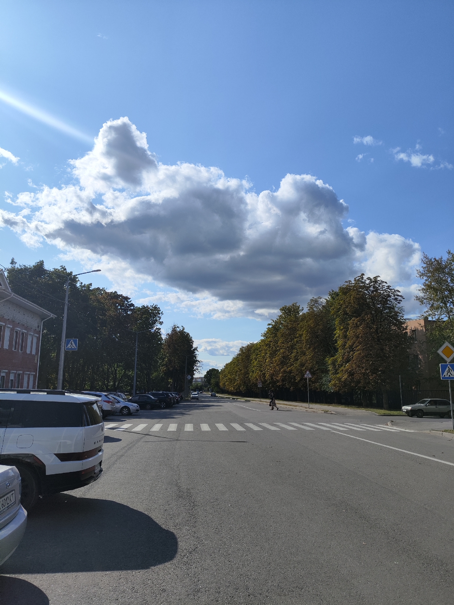 The image depicts a street scene in Kharkiv, Ukraine, on a clear day with scattered clouds. In the foreground, a white SUV is parked on the left side of the road, casting a long shadow. Adjacent to it, a silver car is also parked. The road itself is asphalt, with a pedestrian crossing clearly marked.
Several other cars are parked along the side of the road further down. On the left side of the street, a brick building with multiple windows is visible. Further down the street, a line of trees with autumnal foliage creates a natural border. Streetlights and traffic signs, including a pedestrian crossing sign, are present along the roadside.
In the distance, a few people are visible, including one individual walking across the pedestrian crossing. The sky is predominantly blue, with large, puffy white clouds and some darker, more ominous clouds. The overall scene suggests a typical urban environment on a pleasant day.