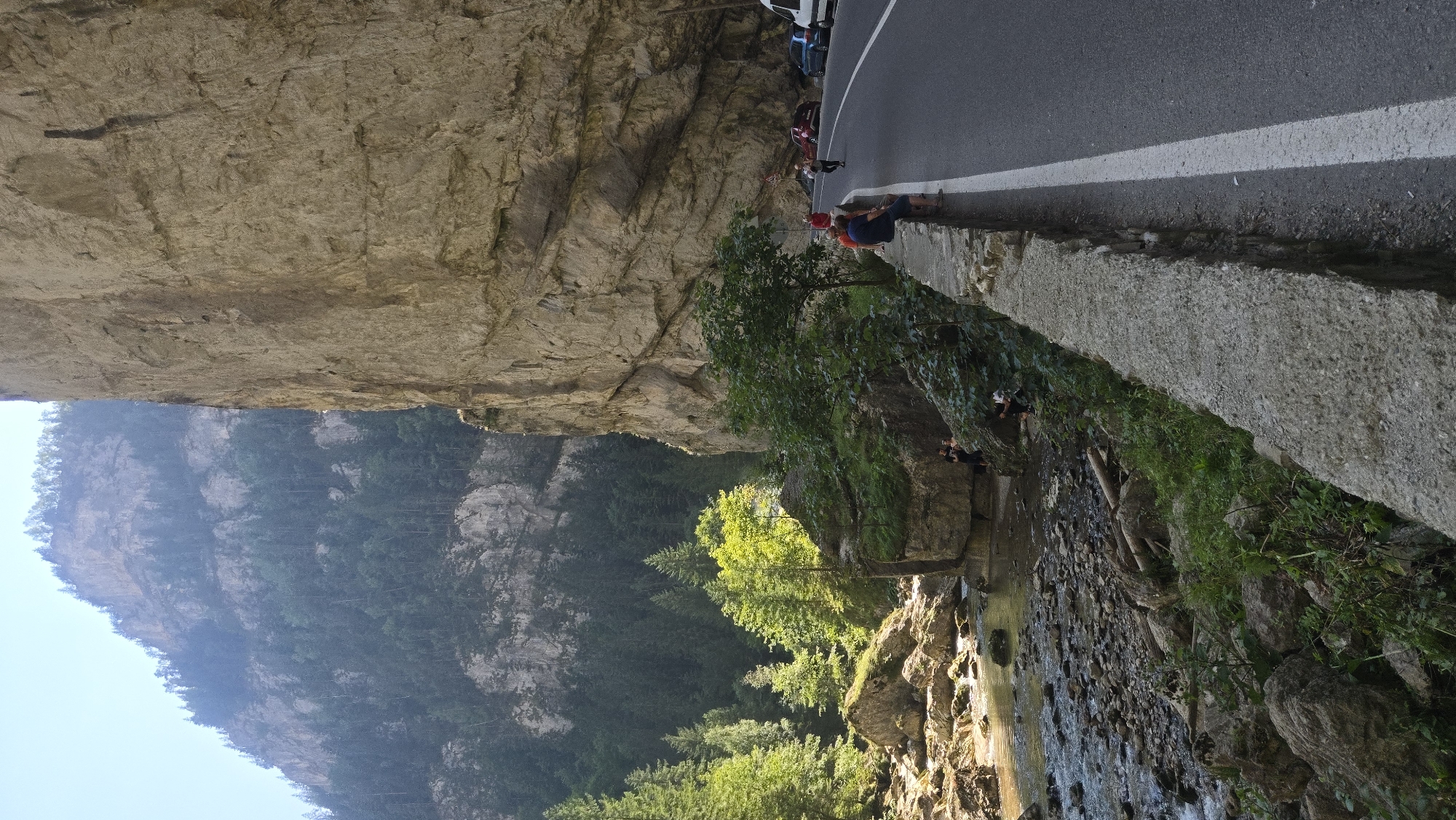 The image depicts a scenic mountain road in Bicaz-Chei, Romania. A winding asphalt road with a white line marking the lane runs alongside a towering, sheer rock face. On the left side of the road, a rocky riverbed with clear, flowing water is visible, bordered by lush green trees and large boulders. Several people are present on the scene. Two individuals are seated on a low stone wall bordering the road, observing the surroundings. Further down the road, a small group of people are gathered near parked cars, including a red car and a blue car. The overall environment suggests a natural tourist attraction, with the mountain scenery and river providing a picturesque backdrop.