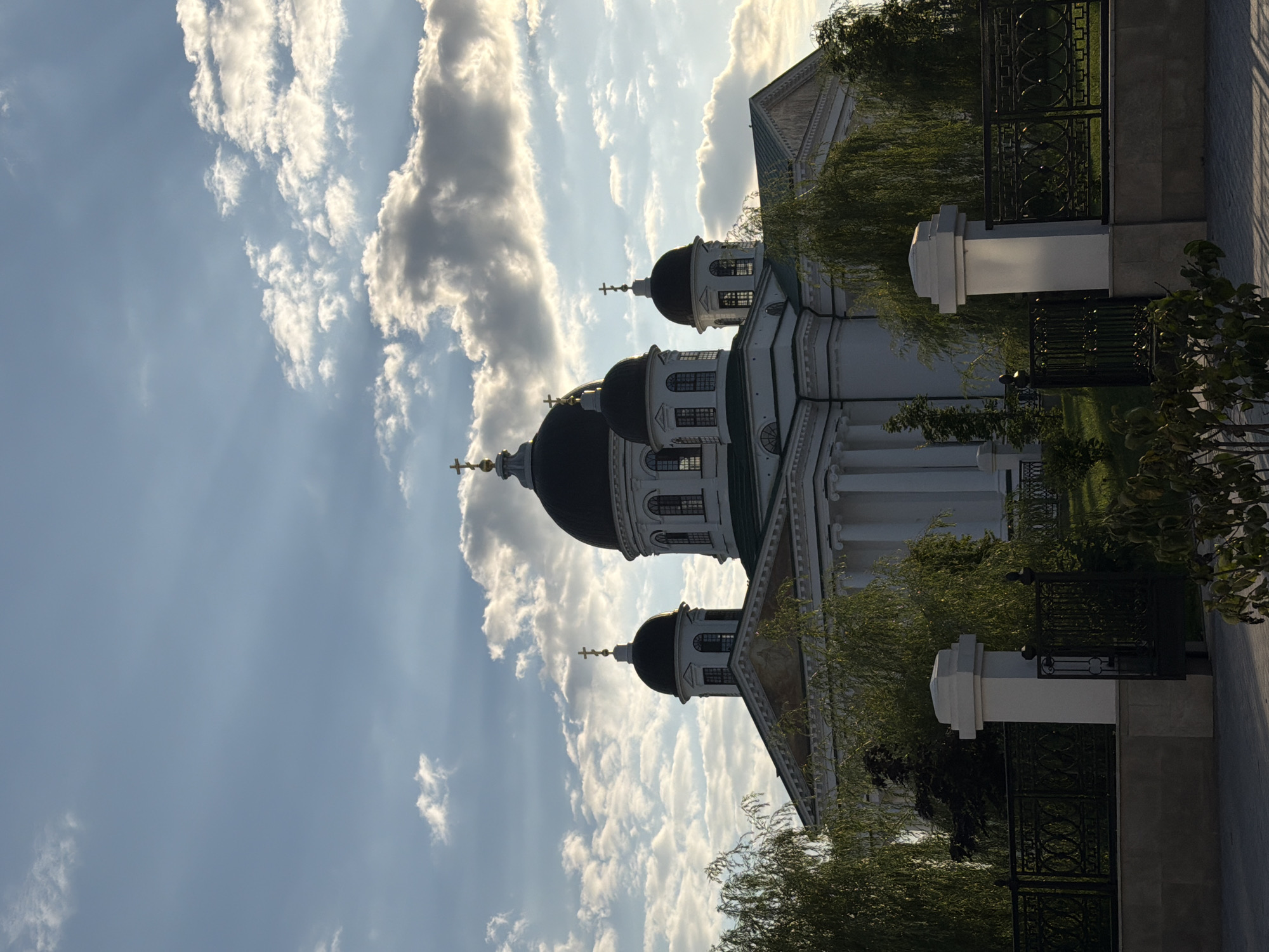 The photograph captures a majestic cathedral with multiple domes in Arzamas, Russia. The cathedral, predominantly white with dark domes, is a prominent structure with classical architectural elements like columns and arched windows. It is set against a backdrop of a dynamic sky filled with scattered clouds, some of which are illuminated by the sun, creating a dramatic effect with rays of light filtering through.
In the foreground, the cathedral is partially obscured by lush green trees, their branches cascading down, adding a touch of nature to the scene. A decorative black metal fence runs across the lower portion of the image, with white stone pillars flanking an entrance, suggesting a controlled access area. The ground in the very front appears to be paved. The lighting suggests it is either late afternoon or early morning, given the angle of the sun and the soft shadows. There are no people visible in the image.