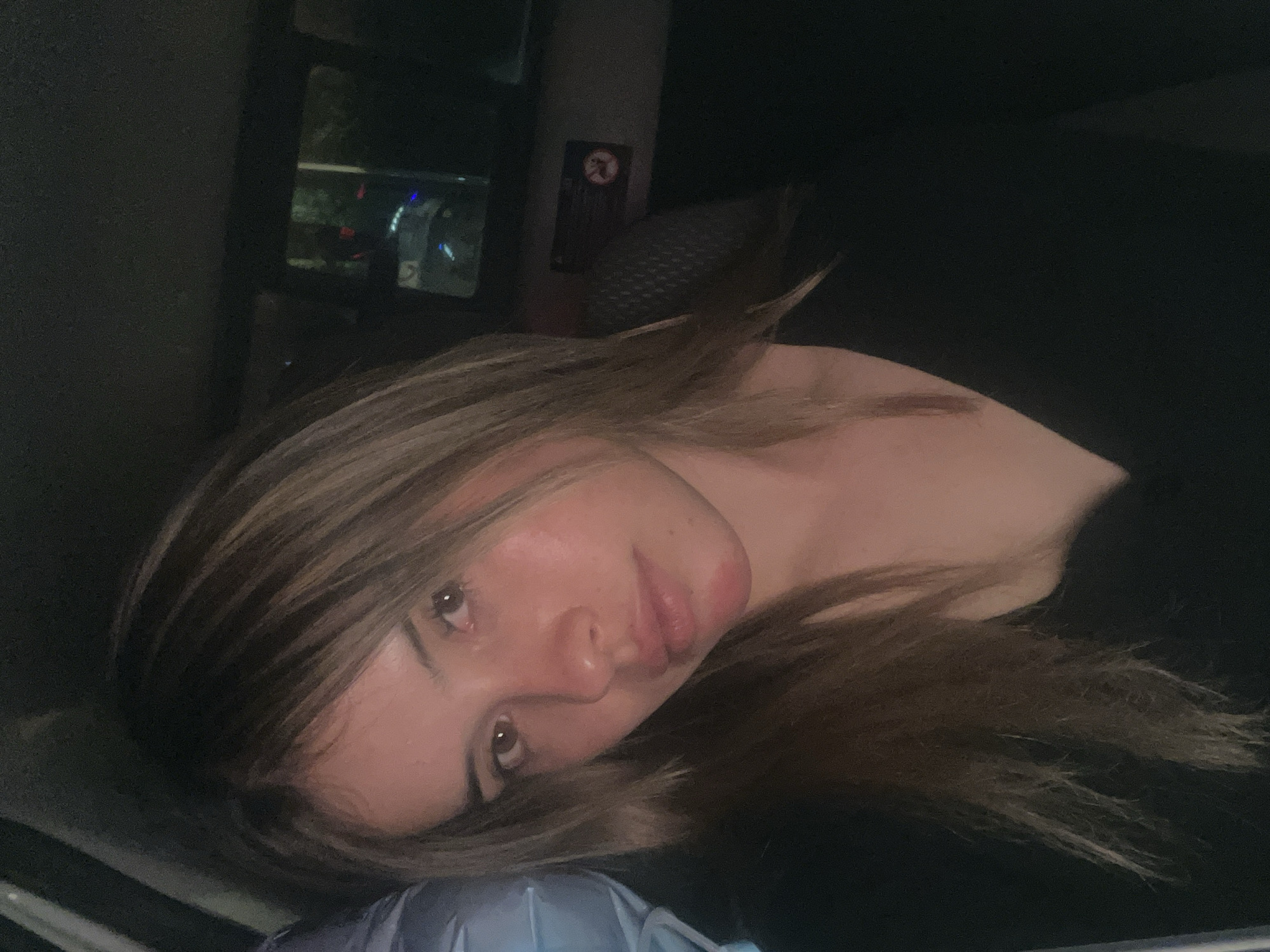 A woman with blonde and brown hair is lying on what appears to be a pillow or soft surface inside a vehicle. The background shows a dark interior with a window reflecting outside lights, possibly from streetlights or other vehicles. A sign is visible above the window, but it is difficult to decipher. The woman is looking towards the camera with a neutral expression. Without additional information, I cannot verify its location in San Fernando del Valle de Catamarca, Argentina.