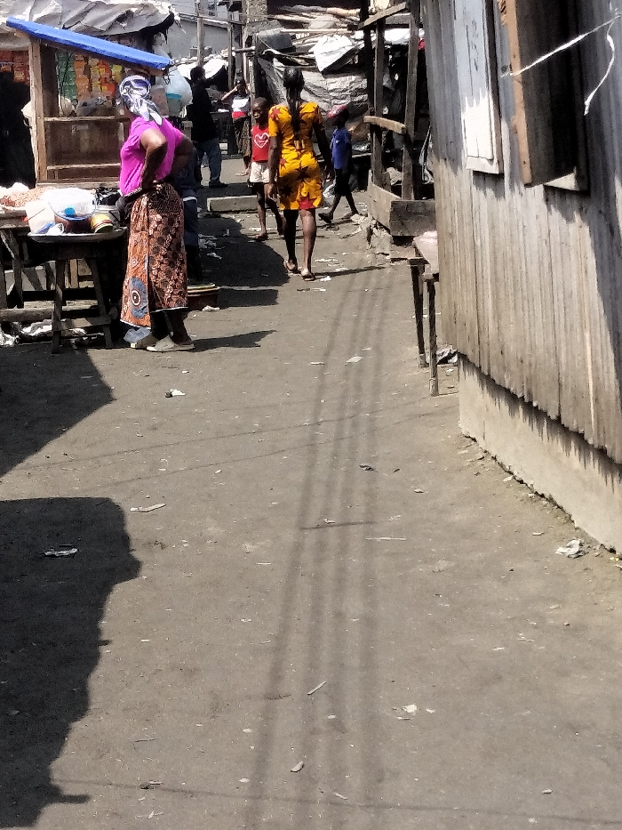The image captures an outdoor scene on an unpaved street or alleyway in Oworonshoki, Nigeria, characterized by informal market activity and residential structures. The ground is dirt-packed, showing scattered debris and refuse. Strong sunlight from an overhead source casts distinct, elongated shadows, including those of individuals and thin linear patterns, possibly from overhead wiring or supports.
On the left, a woman stands with her back largely to the viewer, wearing a vibrant pink short-sleeved top, a multi-patterned brown, orange, and blue wrap skirt, and a white and blue patterned headscarf. She is positioned beside a makeshift wooden stall or table displaying various items, some in plastic containers or bowls, beneath a blue tarp awning.
Further into the scene, centrally located, another woman walks away from the camera, dressed in a yellow and orange patterned short dress. To her immediate left, a young boy in a red shirt walks in the same direction. In the midground and background, multiple individuals are discernible, moving or standing amongst a dense cluster of rudimentary structures, many appearing to be shacks or stalls constructed from corrugated metal sheets and covered with tarpaulins.
The right side of the frame is dominated by the rough, vertically paneled wall of a structure, likely corrugated metal, with a concrete base. A small, open-shuttered window is visible on this wall, and a simple wooden table or stand is positioned in front of it. The overall setting indicates a lively, unformalized urban or semi-urban environment.