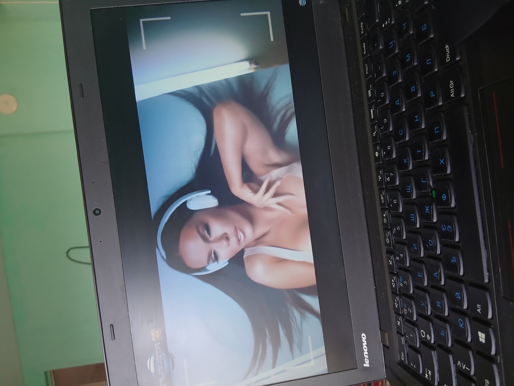 A close-up image shows a black Lenovo laptop with a Cyrillic keyboard. The screen displays an image of a woman wearing headphones and lying down. The laptop is resting on an unidentifiable surface. The environment visible is a blurry green wall. There is no information to determine if this is located in Vetren Dol, Bulgaria.