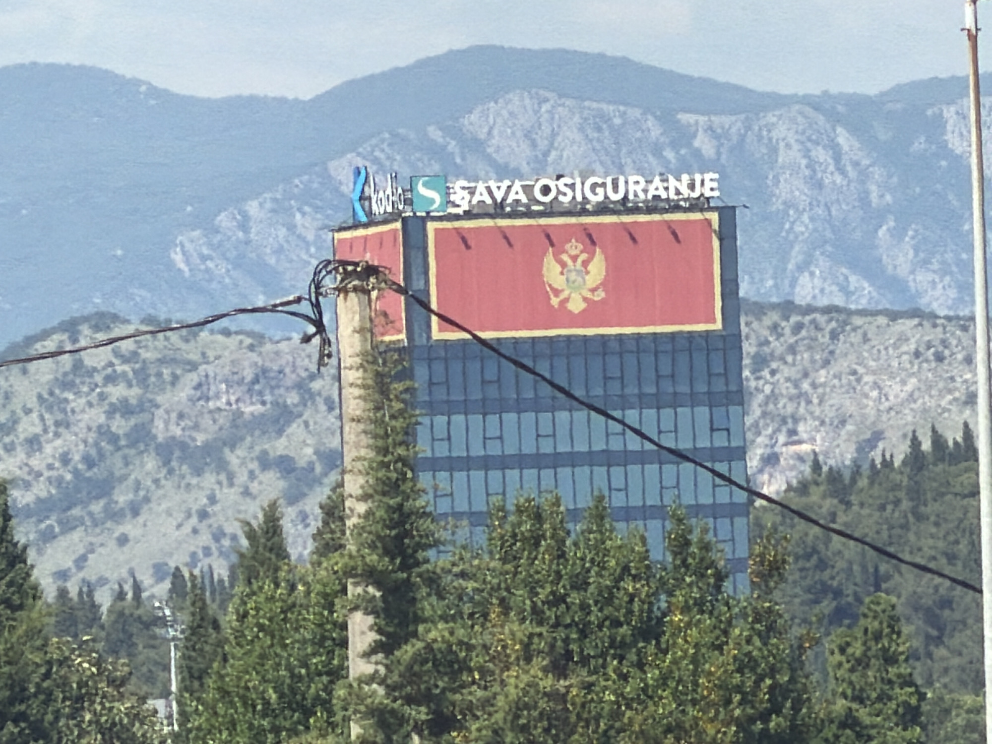 The image shows a tall building in Podgorica, Montenegro. The building has the flag of Montenegro on its upper section, with a golden eagle emblem prominently displayed. Above the flag, the logos of "Kodio" and "SAVA OSIGURANJE" are visible on the rooftop. The building's lower section consists of multiple floors with rectangular windows. In the foreground, trees partially obscure the lower part of the building. Power lines extend across the scene. In the background, mountains are visible. A utility pole is located to the left of the building.